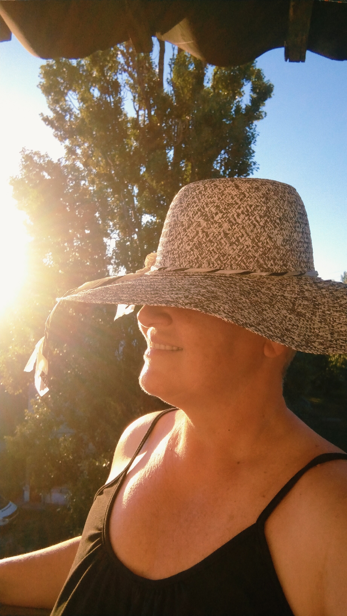 The image shows a person wearing a wide-brimmed hat and a black tank top, seen in profile. The person is looking to their left, with their mouth slightly open, revealing their teeth. The sun is shining brightly from the left side of the frame, creating lens flare and casting strong highlights on the person's face and shoulder. Behind the person, there is a large tree with green foliage against a clear blue sky. The foreground is partially obscured by a dark, curved object, possibly an overhang or roof. The overall impression is that of a person enjoying a sunny day outdoors. No specific location in the city of Horishni Plavni, Ukraine, can be determined from the visual information provided in the image.