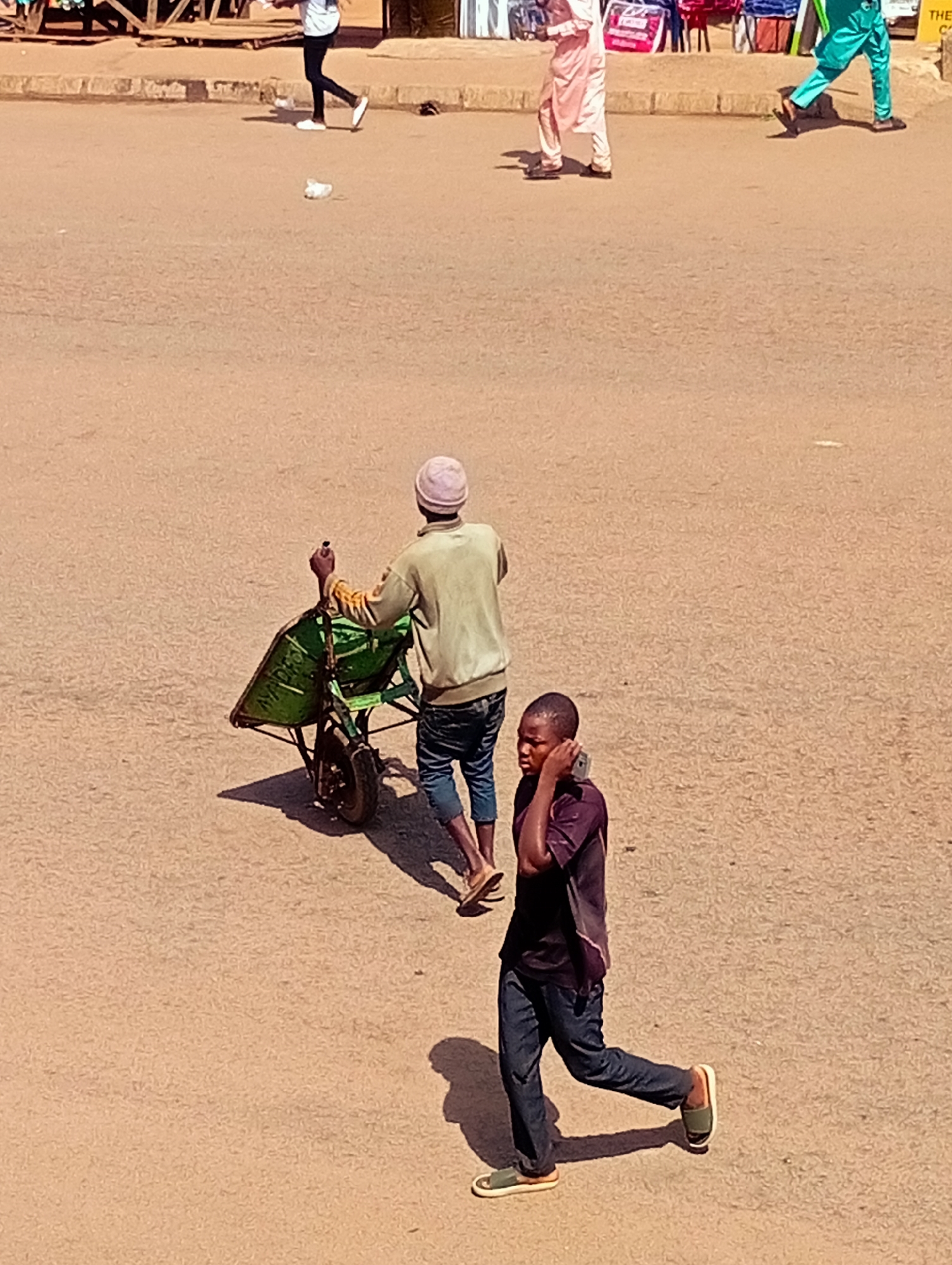 The image depicts an outdoor scene on a sandy or dirt road, likely in Bukuru, Nigeria, during daylight hours. The sun is casting strong shadows, indicating a sunny day. The ground is predominantly a dusty, light brown.
In the foreground, a young Black male is walking towards the right, with his right hand raised to his ear as if speaking on a phone. He is wearing a dark purple short-sleeved shirt and dark blue jeans. He has on dark sandals with light-colored soles. His gait suggests he is in motion.
Behind him and slightly to the left, an individual wearing a light pink cap is pushing a green wheelbarrow. This person is dressed in a light-colored long-sleeved shirt and dark pants. The wheelbarrow appears to be loaded.
Further in the background, other individuals are visible. To the far left, a person in white and black is walking away from the camera. To the right, a person in bright teal pants and a lighter top is walking towards the right. A few market stalls or structures are visible along the upper edge of the frame, with various colored bags or merchandise. Some white debris is scattered on the ground.
The overall scene suggests a busy street or marketplace environment, with people going about their daily activities, possibly involving transport of goods, indicated by the wheelbarrow. The activity appears to be ordinary daily life.