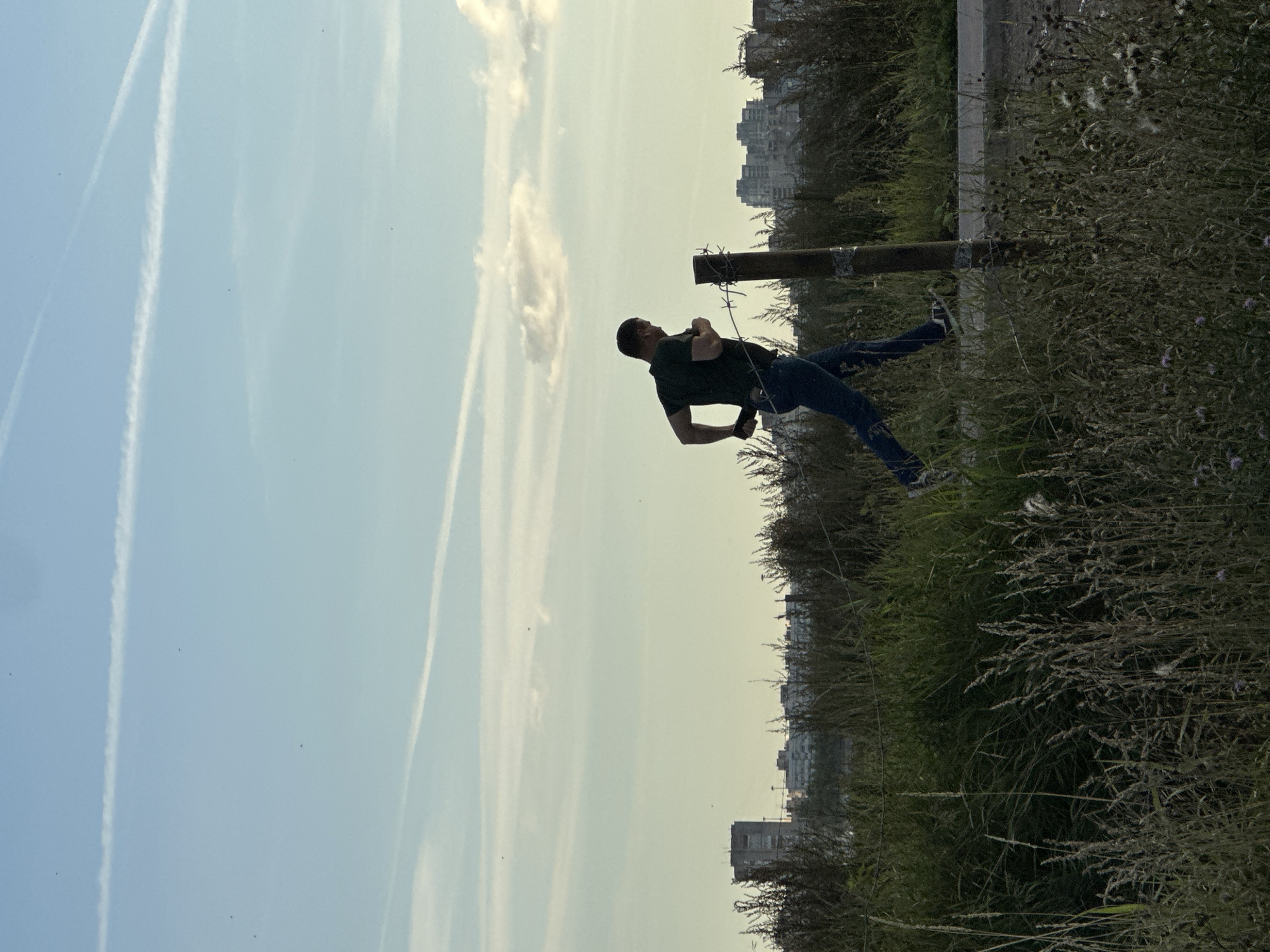 A man is jogging past a barbed wire fence in a grassy, undeveloped area. He is wearing a dark t-shirt, blue jeans, and sneakers. The fence post is made of wood and wrapped with wire. In the background, buildings and apartment complexes are visible, suggesting an urban or suburban environment. The sky is light blue with thin white clouds and contrails from airplanes. The time of day appears to be late afternoon or early evening, indicated by the soft lighting. The location is the outskirts of Murino, Russia.