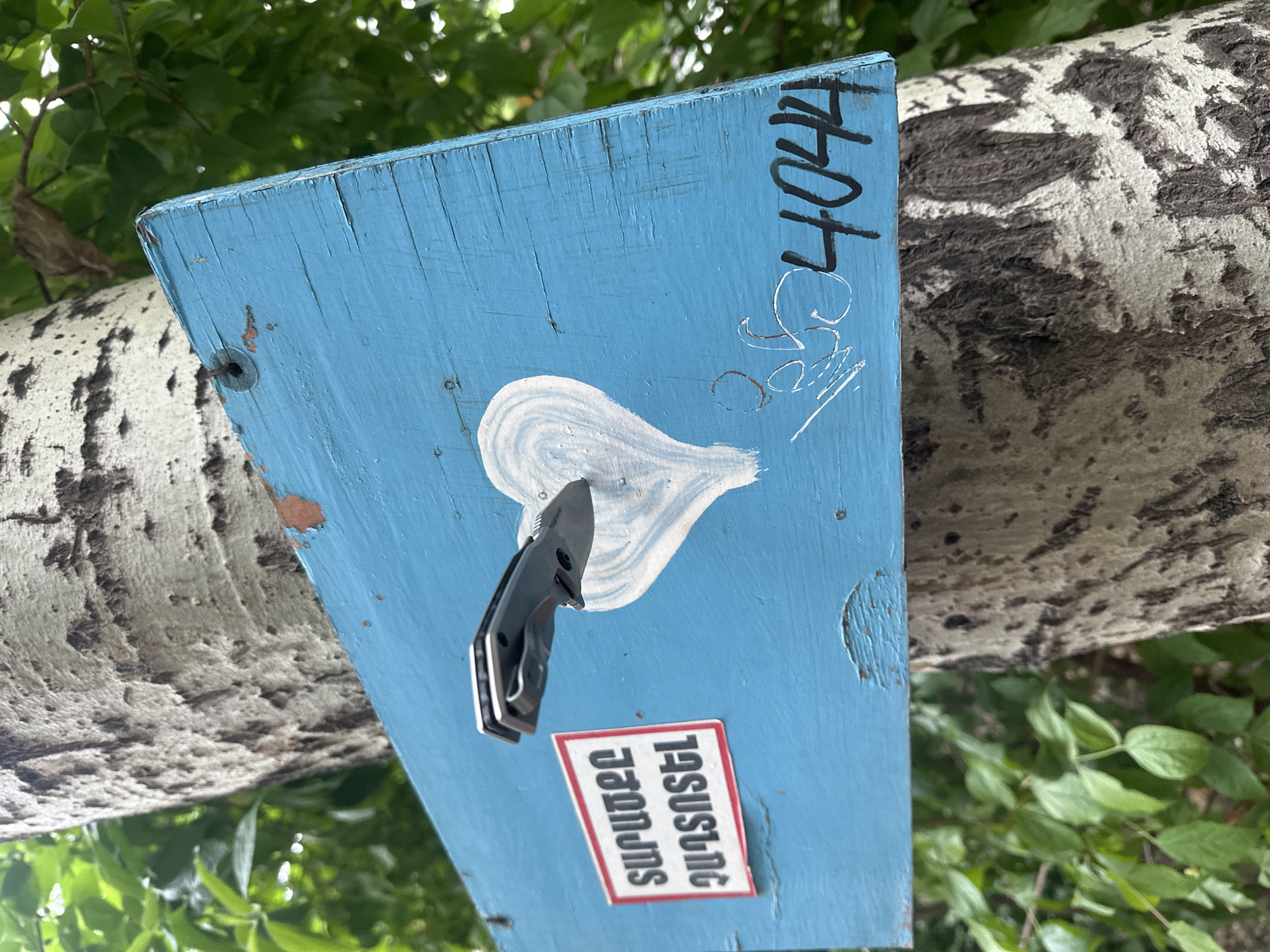A blue wooden sign is attached to a tree, possibly in Yerevan, Armenia. The sign features a white painted design resembling a teardrop or heart, and has a black folding knife attached to it. The number "4044" is written in black marker along the top edge of the sign. Below the knife, there is a sticker with Armenian text on a red background. The tree bark is textured and grey. Green foliage is visible in the background.