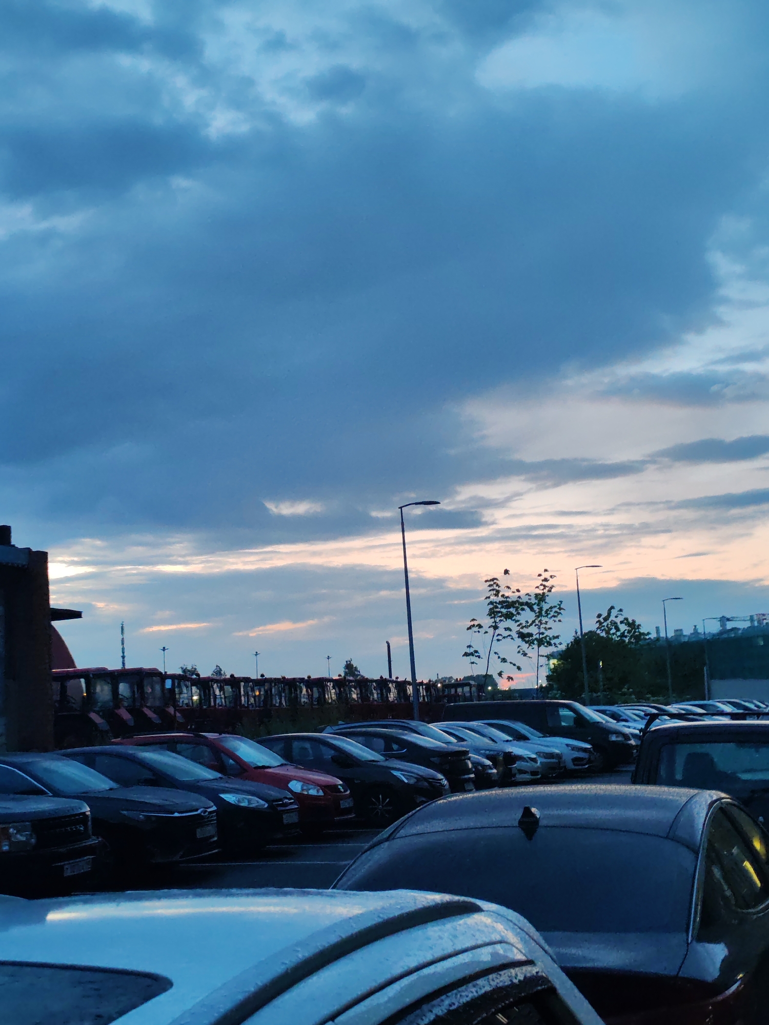The image shows a parking lot with several cars parked in rows. In the background, a line of red tractors is visible, along with trees, streetlights, and distant buildings. The sky is overcast with clouds, and there are hints of sunset colors in the distance. The scene appears to be in Shushary, Russia, based on the provided context. There are no people visible in the image, and no specific actions or interactions are depicted. The environment is that of an outdoor parking area with a mix of vehicles and infrastructure under a cloudy sky.