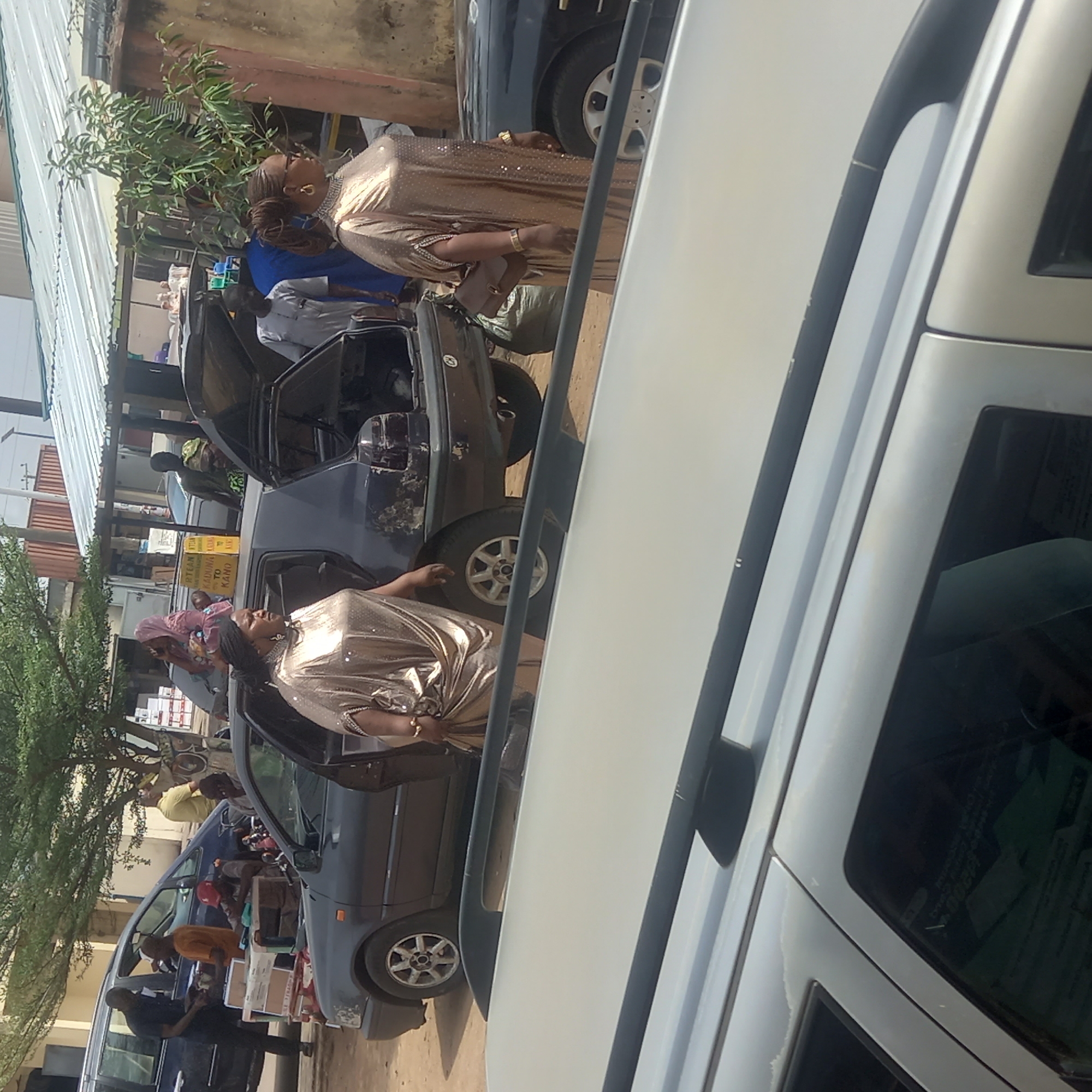In Dadi Riba, Nigeria, a street scene unfolds with multiple vehicles and pedestrians. A woman in a shimmering gold dress stands near a dark-colored vehicle with its door open. Several people are engaged in activities around other cars, including what appears to be loading items. Buildings are visible in the background, along with trees and foliage. A white vehicle is prominently featured in the foreground.