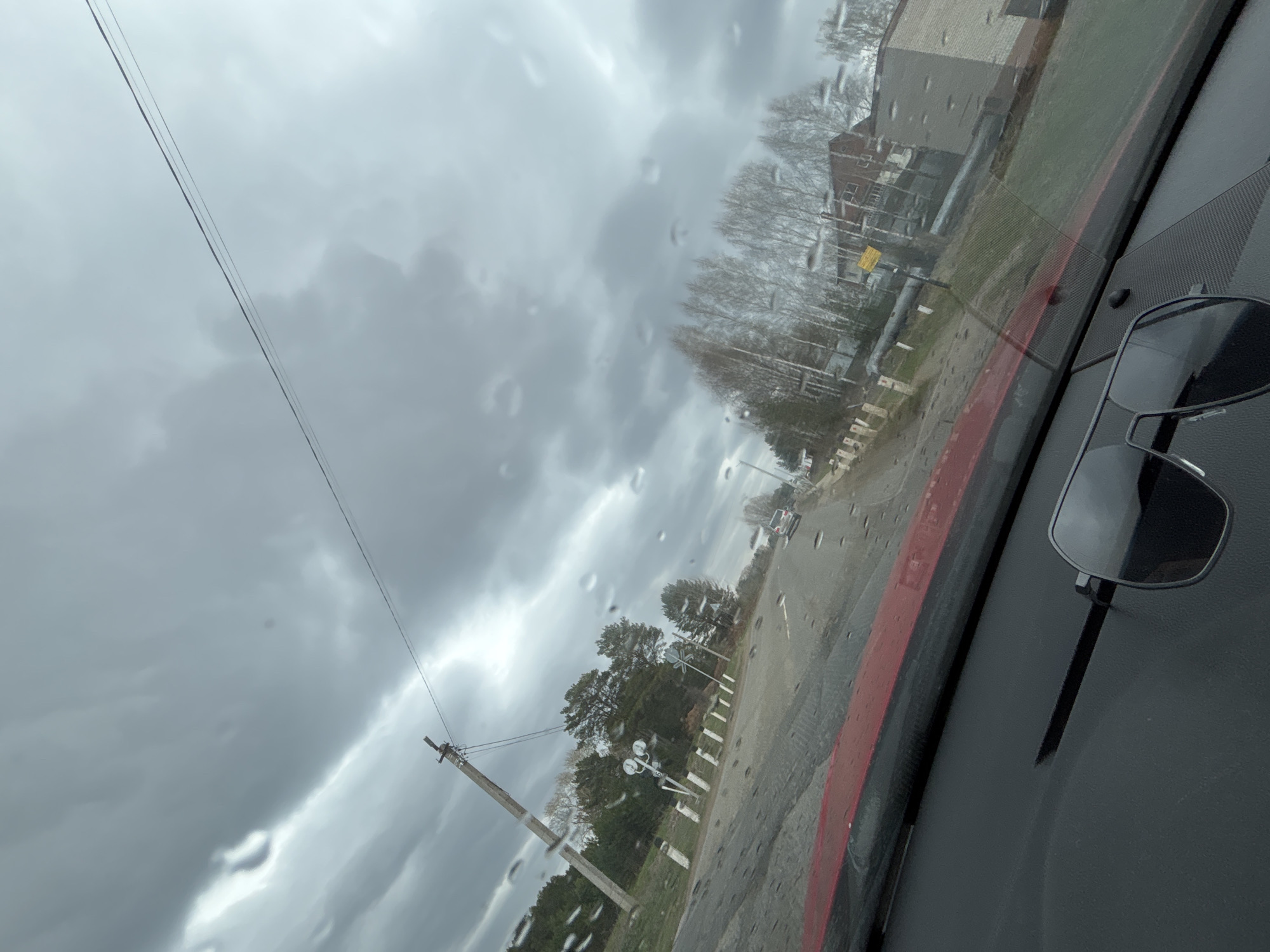 The image captures a gloomy, overcast daytime scene from the interior of a vehicle in Chaikovsky, Russia, looking out through a rain-streaked windshield. Dark, heavy clouds fill most of the sky, suggesting recent or ongoing precipitation.
Thin black power lines stretch across the upper left portion of the sky, connected to a tall, wooden utility pole in the mid-ground. To the right of this pole, two white "crossbuck" railroad crossing signs are visible, with the word "RAILROAD" partially legible on one. A road or track runs from the foreground into the distance, lined with white marker posts. Beyond the road and tracks, a mix of bare deciduous trees and some evergreen foliage is present, indicative of late autumn, winter, or early spring. In the background, partially obscured by trees, some buildings are visible, including one with reddish-brown brick. A faint outline of another light-colored vehicle can be seen further down the road in the distance.
The bottom right of the image shows a segment of the vehicle's dark dashboard, which has a textured surface and a distinctive red trim running along the base of the windshield. A pair of dark-lensed sunglasses rests on the dashboard. The raindrops on the windshield confirm the wet weather conditions, contributing to the subdued and somber atmosphere of the scene.