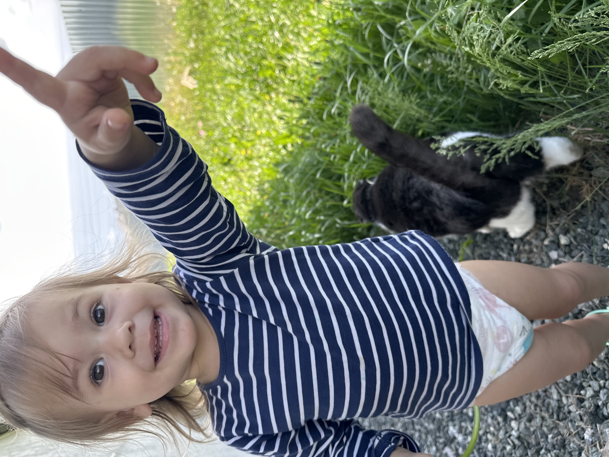 A young child in a blue and white striped shirt and diaper is standing outside on gravel. The child has their right arm raised and is smiling at the camera. Beside the child, in the grass, is a black and white cat. The environment appears to be a yard or outdoor space. No identifiable landmarks to place the location within Kharnavy, Russia are visible.