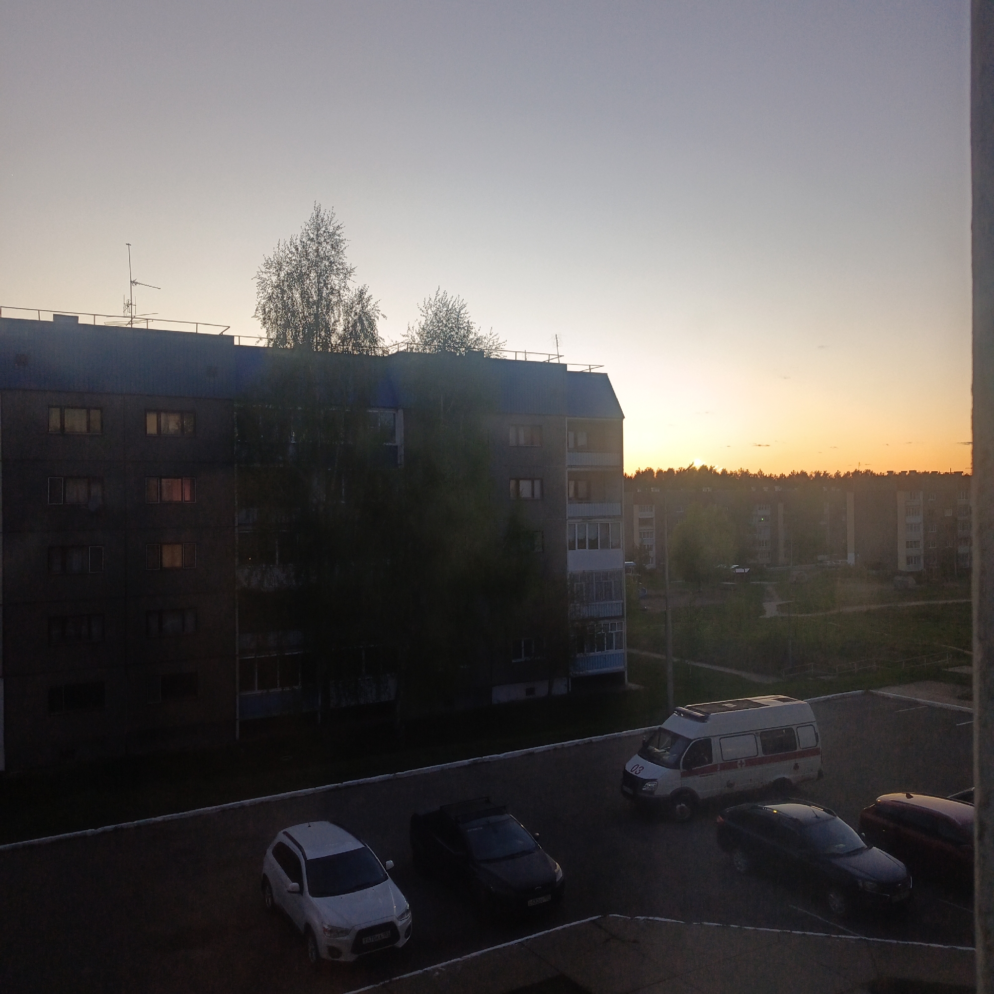The image shows an urban scene at sunset in Марковский, Russia. In the foreground are several parked cars and an ambulance in a parking lot. Beyond the parking lot, there is a multi-story apartment building partially obscured by trees. In the background, there are more residential buildings and a treeline silhouetted against the colorful sunset sky. The image is taken from an elevated viewpoint, likely from a window or balcony.