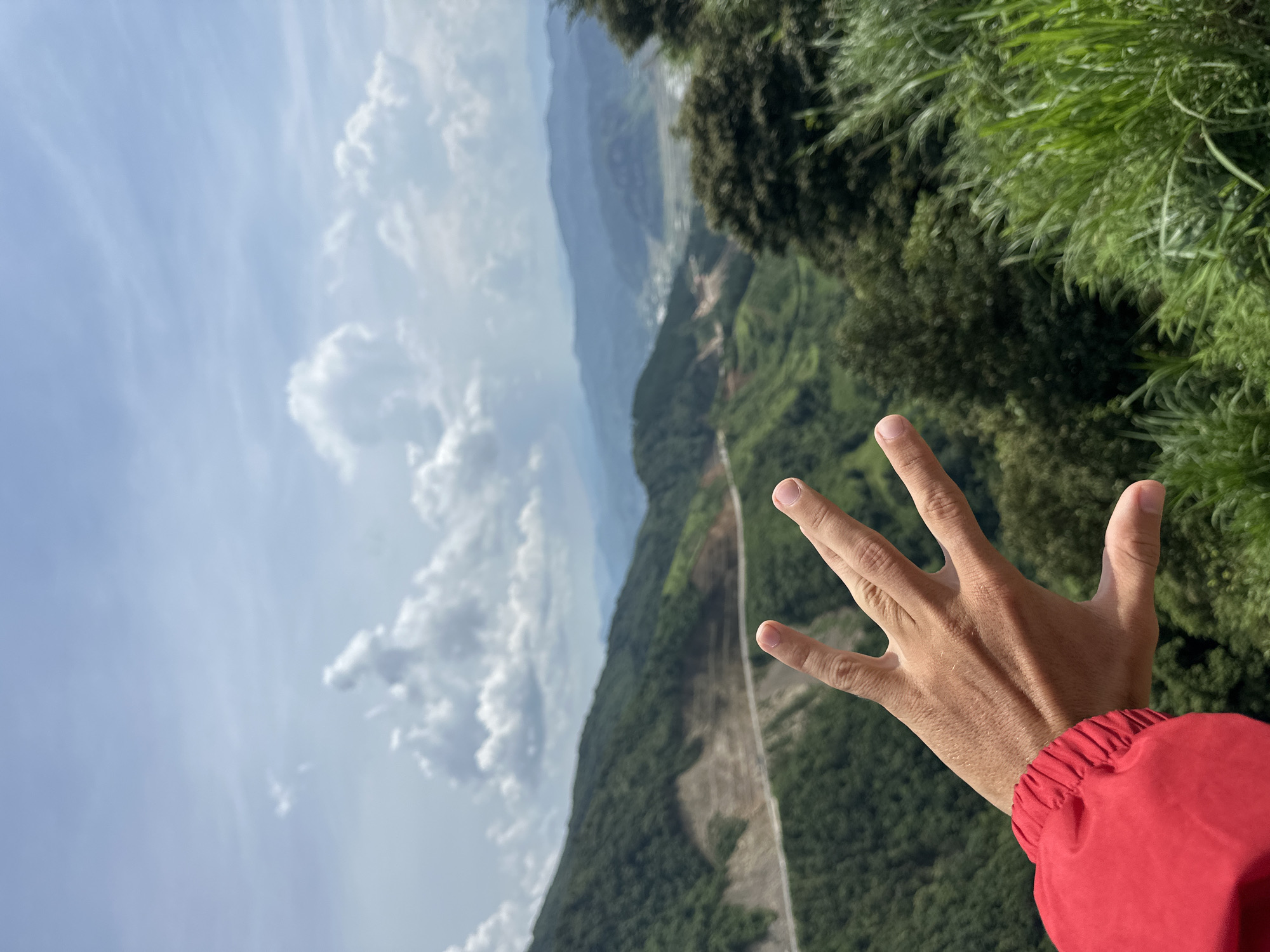 A vibrant, daytime, outdoor shot captures a panoramic view of a mountainous landscape from a high vantage point, with a person's right hand prominently featured in the foreground. The hand is open, fingers slightly spread, and appears to be held out towards the expansive vista. A red jacket sleeve with an elastic cuff is visible on the wrist.
The background reveals a vast valley carved between numerous layers of green-clad mountains and hills that recede into a hazy, blue distance. A distinctive light-colored road, possibly unpaved in sections, snakes and winds through the verdant terrain, traversing the valley floor and ascending the slopes. Evidence of land disturbances, perhaps from construction or agricultural use, can be seen along the road and hillsides. The immediate foreground on the right side features lush, bright green foliage and tall grasses.
The sky is a clear, bright blue, adorned with scattered white clouds, ranging from wispy cirrus to more defined cumulus formations. The lighting suggests it is a sunny day, likely mid-morning or afternoon, offering excellent visibility of the distant topography. The overall scene conveys a sense of awe and scale, consistent with a high mountain overlook in a location like Muong Than, Vietnam. No discernible text is present in the image.
