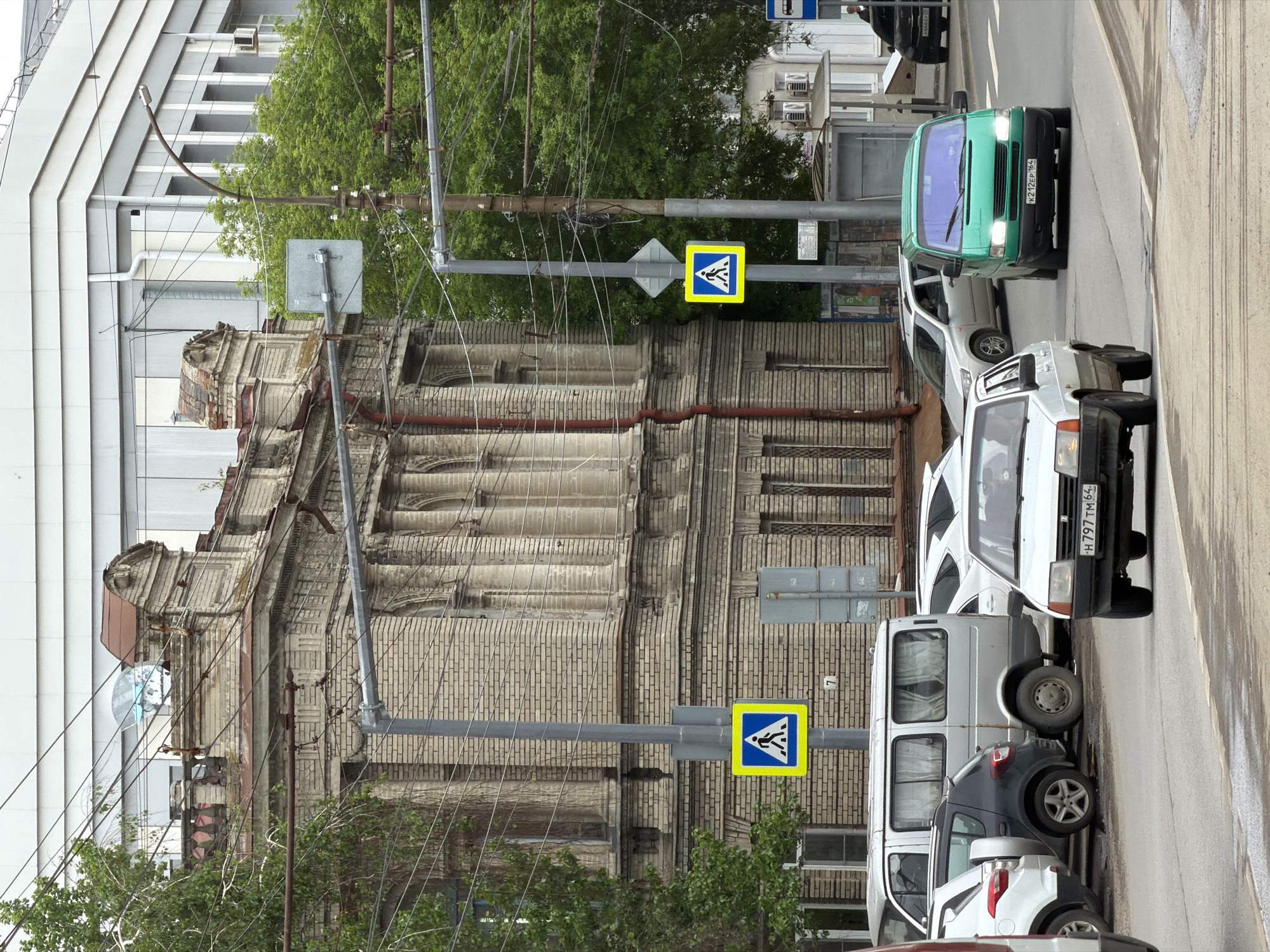 The image captures a street scene in Saratov, Russia, with an older brick building situated next to a modern white building. A pedestrian crossing sign stands in front of the brick building. Parked vehicles line the side of the road, including a green van with the license plate "K212EP164" and a white car with the license plate "H797TM64." Overhanging wires and sparse greenery are visible near the building and traffic signs.