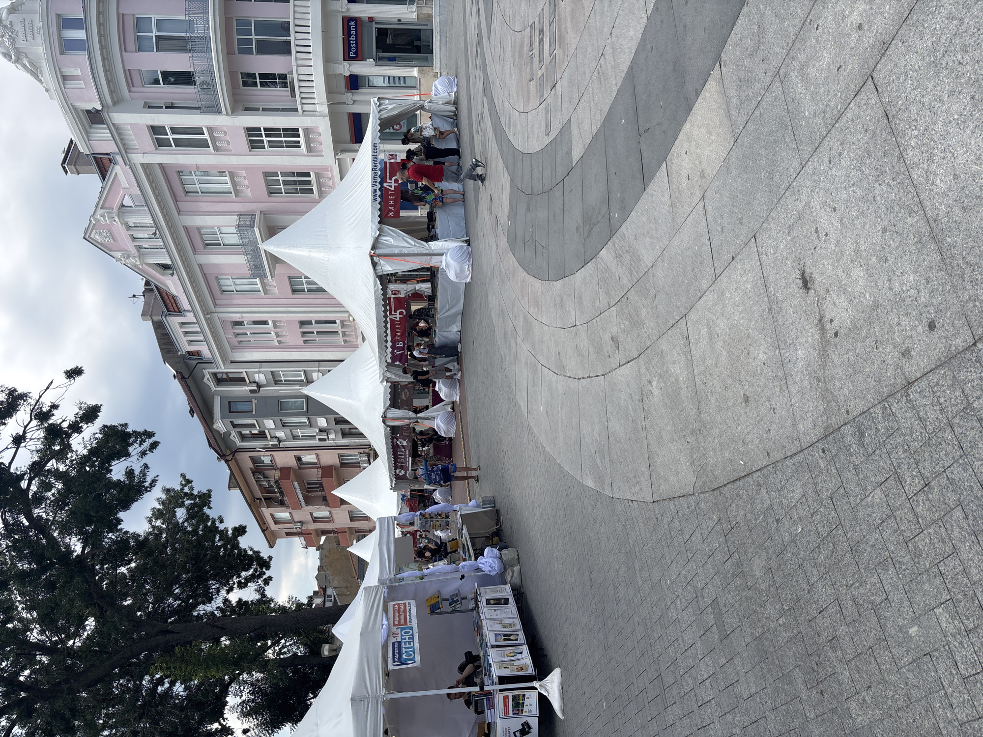 This is a street-level, eye-level, medium shot photograph taken in Varna, Bulgaria, featuring a temporary outdoor market or exhibition. The scene is dominated by white tents set up in a paved public square. In the foreground, a large tree with dark green foliage on the left casts shadows. The tents are arranged in a row, with several visible, including one on the far left displaying books and promotional materials for "IZDATELSTVO STENO" and "STENOBIOKS.COM." A person is seated behind a table within this tent.
Further down the row of tents, more white structures are present, some with visible signage. One tent has a banner with "www.VarnaRental.com" and "ЖАНЕТ 45" displayed on it. People are seen walking around and interacting within and around the tents. In the background, several multi-story buildings line the square, with architectural styles indicating an urban setting. The building on the far right is painted pink and white, with a "Postbank" sign visible on its facade. The pavement is made of gray stone tiles, laid out in a patterned design with curved pathways. The sky is overcast.