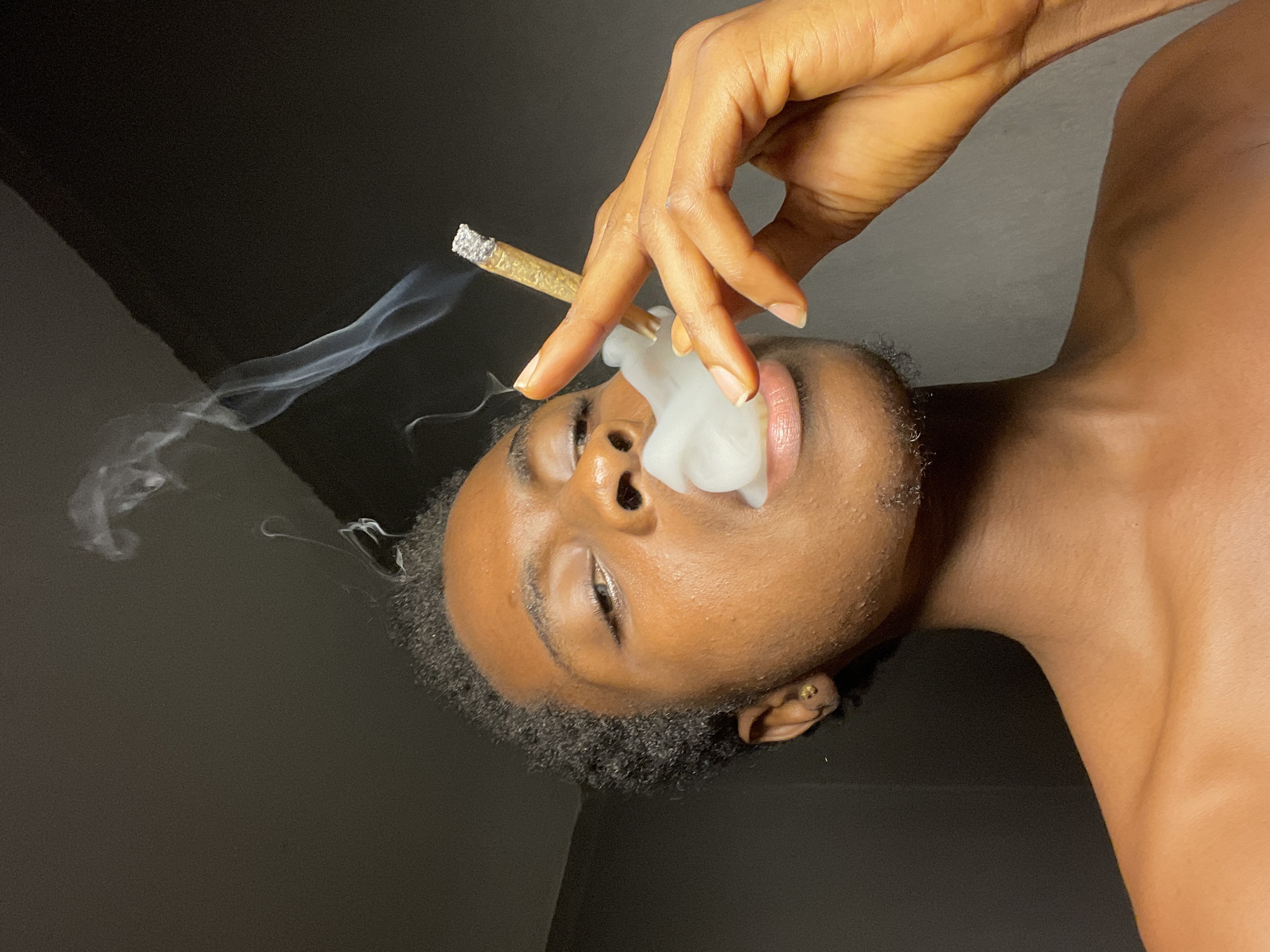 A person is holding a lit marijuana cigarette and exhaling smoke. The person has brown skin, short black curly hair, and a gold earring. The background is dark. The location is unknown.