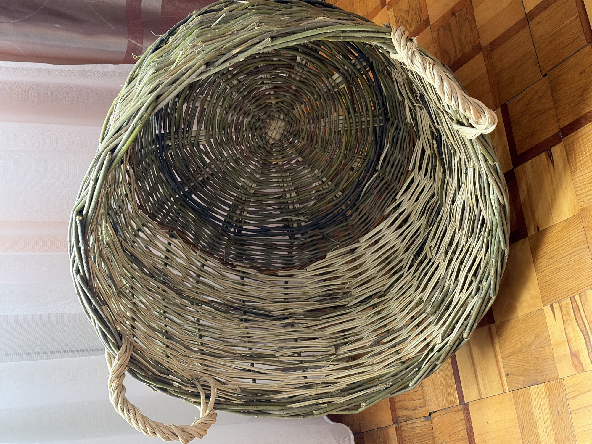 A large, round wicker basket sits on a polished wooden floor. The basket is made of natural materials, with a woven pattern that transitions from light tan to dark green. It has two thick, twisted rope handles, one on each side. The basket appears to be empty. In the background, sheer white curtains hang against a wall. The overall impression is of a rustic, handcrafted item in a home environment.