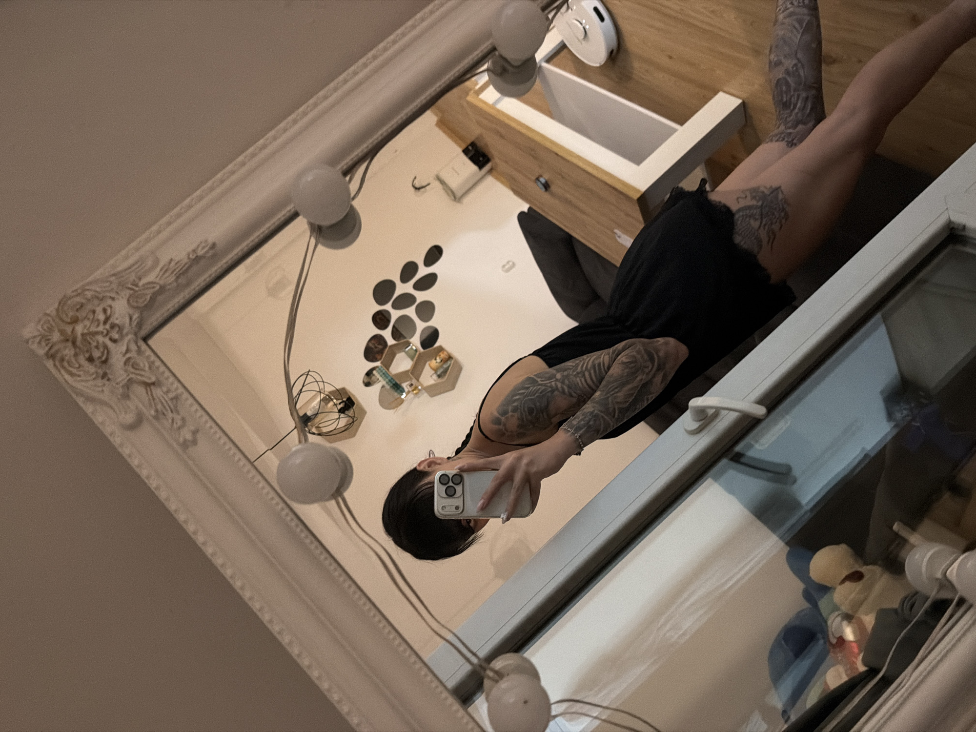 A person with extensive arm and leg tattoos is taking a mirror selfie. They are wearing a black dress and holding a smartphone. The mirror has an ornate white frame. In the reflection, a wooden table, a sofa, wall decorations, and light fixtures are visible. The scene appears to be indoors, likely a living room or bedroom. The image does not provide any information about the location being in the city of Plovdiv, Bulgaria.