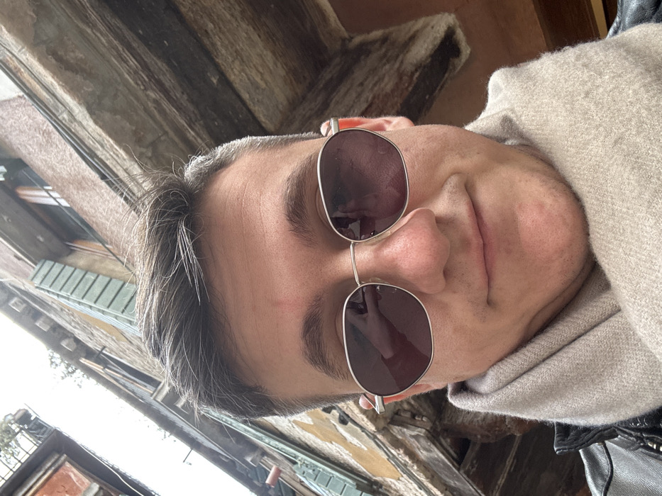 A young adult male, with short brown and grey hair, wears silver-framed, dark-lensed sunglasses, a beige knitted scarf, and a black leather jacket. He is positioned facing the camera, exhibiting a slight smile. A faint red mark is visible on his forehead above his left eyebrow. The background reveals a narrow urban setting characteristic of Venice, Italy, featuring tall, weathered stone and stucco buildings in shades of brown, beige, and reddish-orange. Visible architectural details include a window with closed light green wooden shutters on the left, an open window frame on the right, and exposed wooden support beams under the building eaves. An overcast sky is visible in the gap between the buildings. Sparse green foliage appears on an upper ledge, and dark cables run across a building facade.