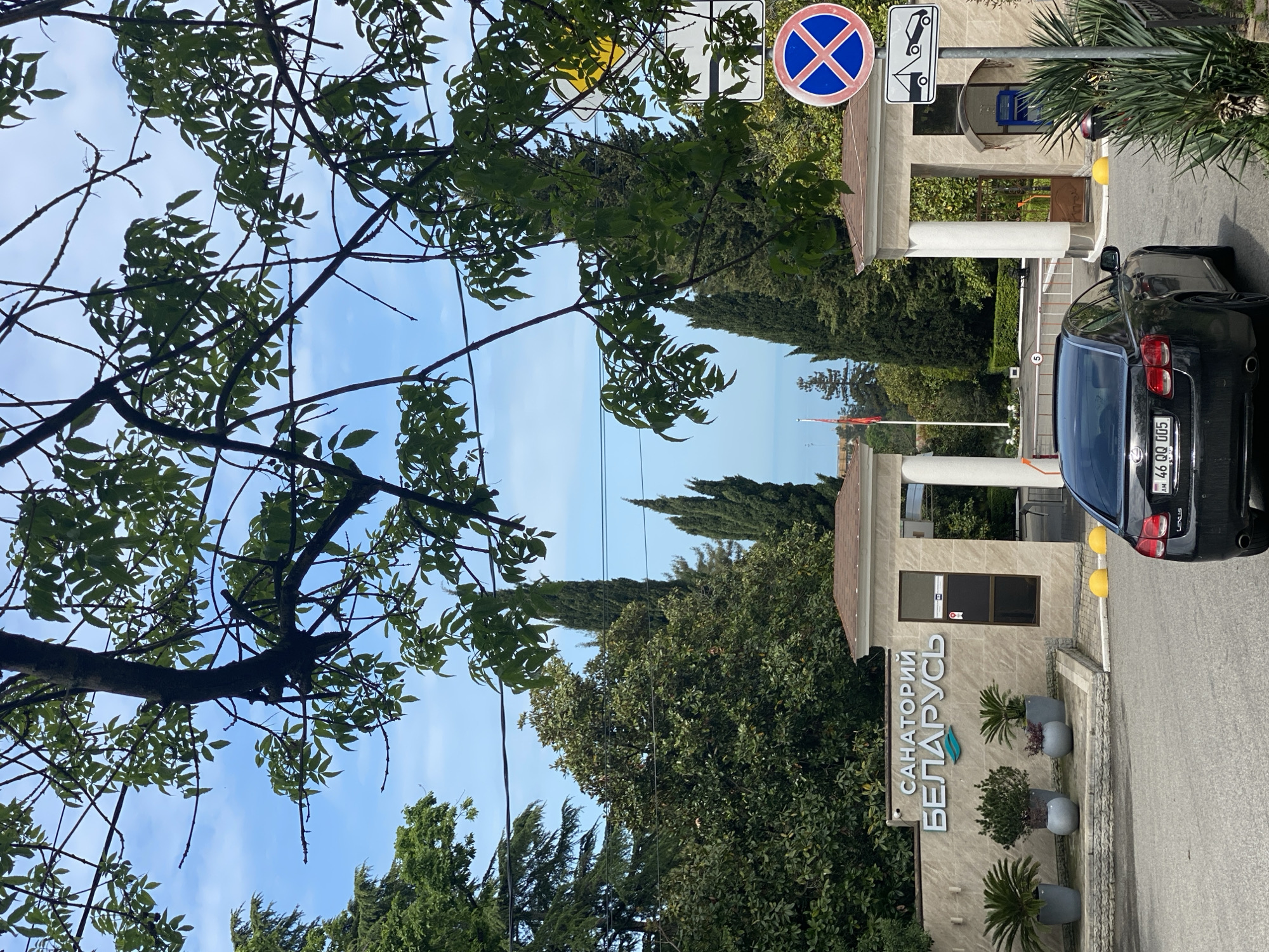 The image shows the entrance to the "Sanatorium Belarus" in Sochi, Russia. A black car is parked near the entrance gate, next to a sign indicating "No Stopping" with a tow-away zone. The building has a tiled roof with beige pillars and signage in Russian "САНАТОРИЙ БЕЛАРУСЬ". Potted plants are placed along the entrance. The scene is partly shaded by trees and shows a blue sky.