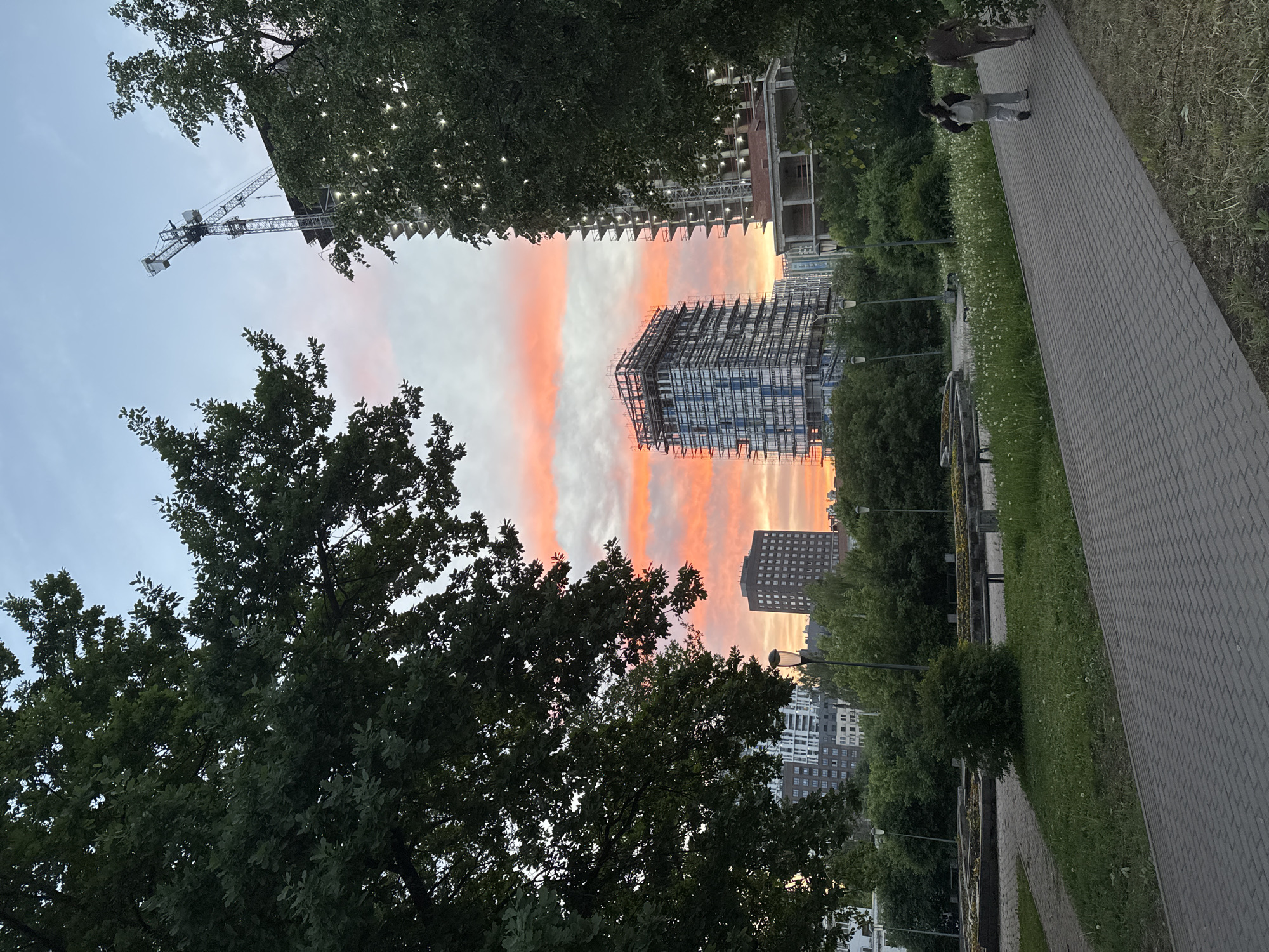 The image shows a view in what appears to be Izhevsk, Russia. The time of day is likely late evening or early morning, judging by the sky with its pink and orange-hued clouds, which could indicate either sunrise or sunset.
Several buildings are in the process of construction, as evidenced by cranes and scaffolding. These unfinished buildings stand amidst a backdrop of other, more completed city buildings. Trees are prominent in the foreground and the middle ground, framing the cityscape. A brick walkway is visible on the right-hand side, with a grassy area adjacent to it. Two individuals are walking along the walkway.
The image provides a juxtaposition of natural and urban elements, with construction sites, the urban park, and people moving along the walkway.
