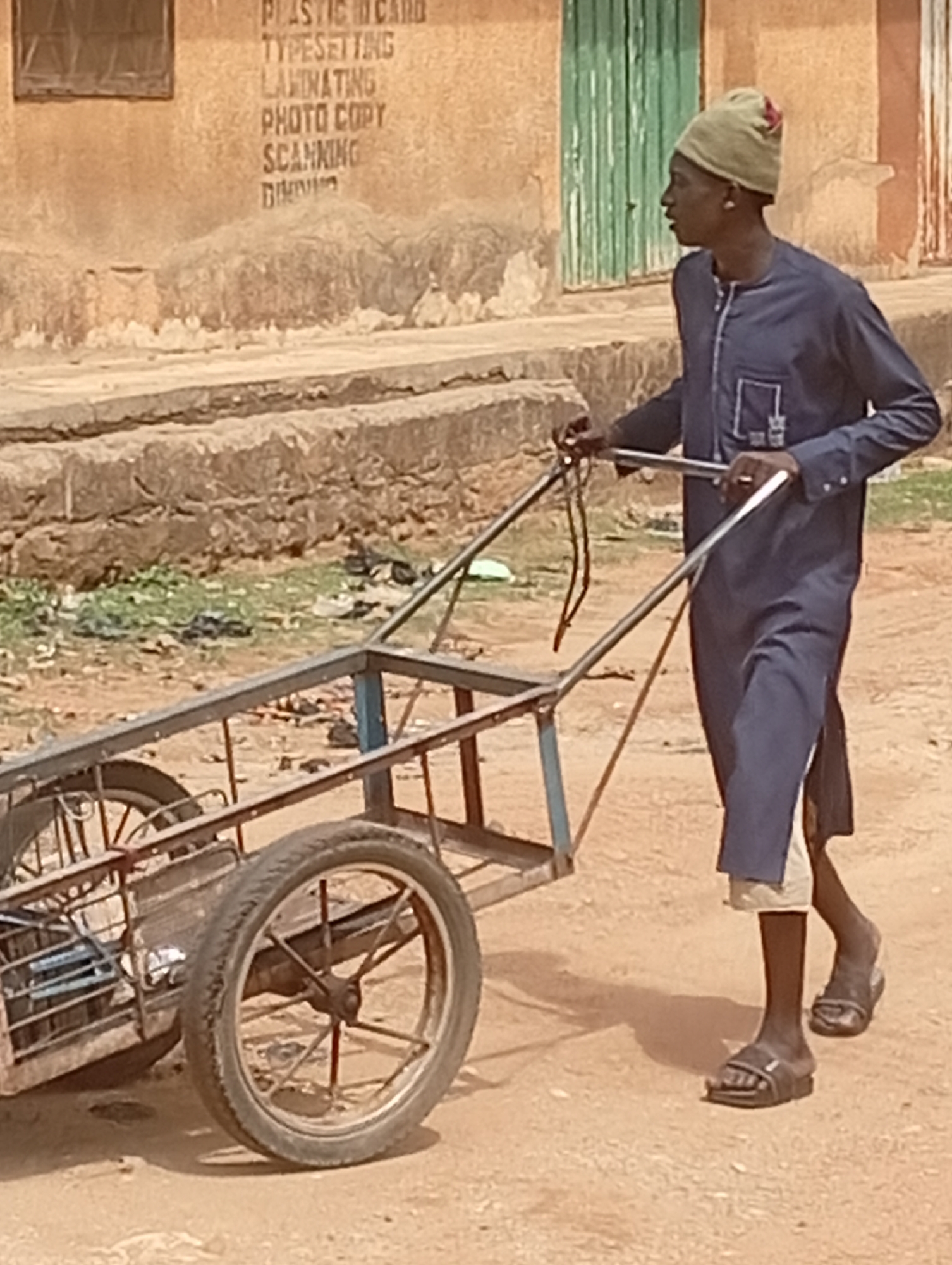 In Sabon Tasha, Nigeria, a Black man in a long navy blue tunic, light beige undershirt and pants, and brown sandals pushes a metal-framed, two-wheeled cart. He also wears a light green knit hat with a red accent. The ground is dry and dusty. In the background is a tan-colored building with black text listing "PLASTIC ID CARD," "TYPESETTING," "LAMINATING," "PHOTO COPY," and "SCANNING." A green door is also visible. The scene is outdoors in daylight.