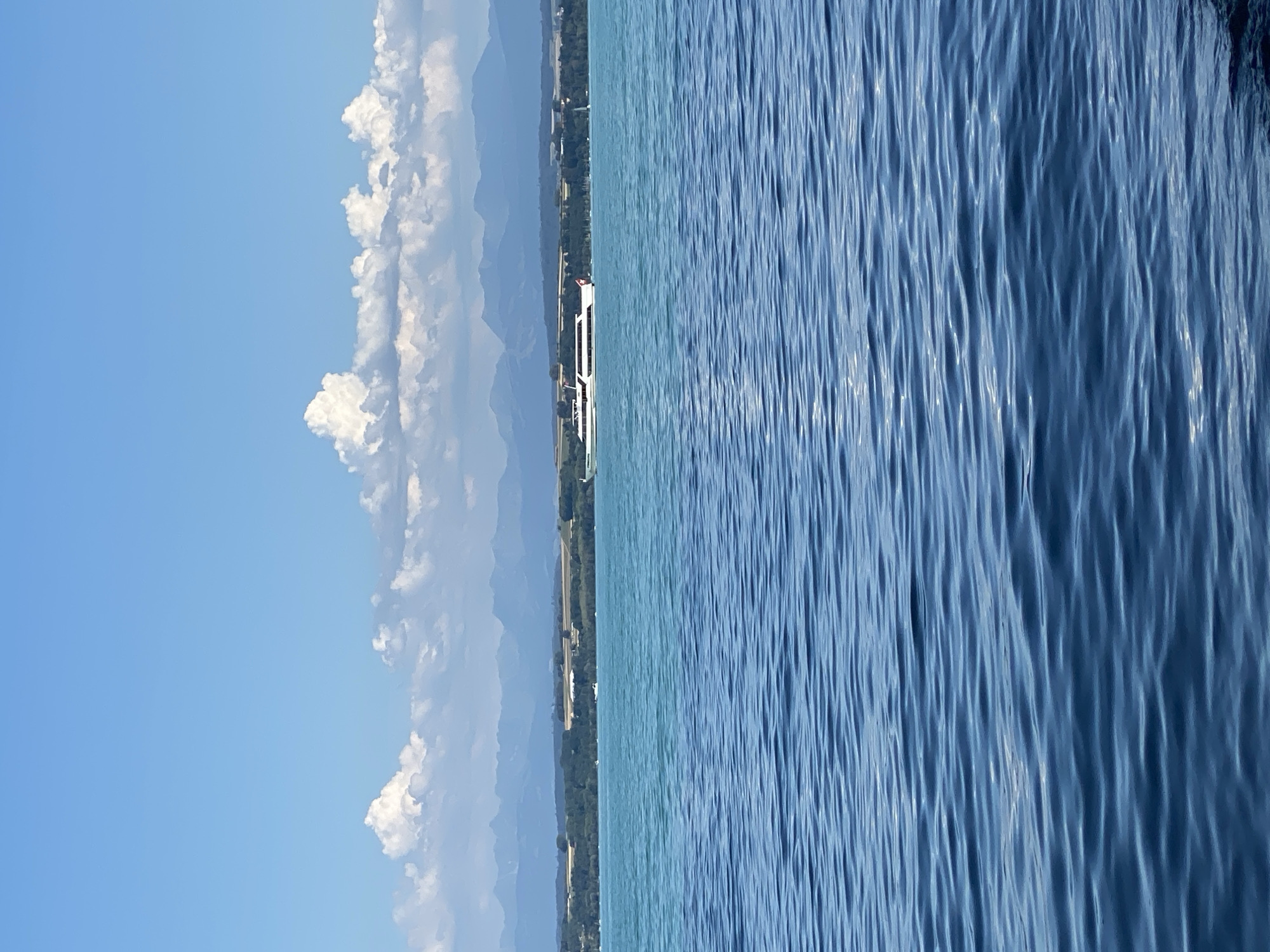 The image depicts a bright, sunny day over a large body of water, likely a lake. The foreground is dominated by the deep blue, rippling surface of the water. In the midground, a distinct turquoise band of water separates the foreground from the far shore. Along the far shore, a tree-lined bank is visible, with fields and buildings beyond. In the distance, a mountain range with hazy blue peaks stretches across the horizon under a bright blue sky filled with fluffy white clouds. A white passenger boat with multiple decks is cruising on the turquoise water, parallel to the shore. The scene appears to be a serene and picturesque landscape, typical of a lakeside environment in a temperate climate. The specific location is the city of Cortaillod, Switzerland, known for its position on the shores of Lake Neuchâtel.