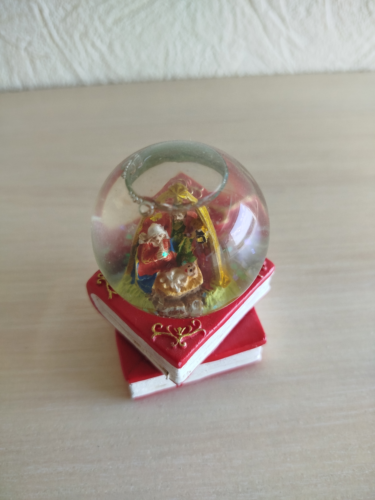 A close-up, eye-level shot shows a small nativity scene inside a glass orb, which rests on two stacked, red, open books. The nativity scene features miniature figures of Mary, Joseph, and a baby Jesus in a manger, with a manger backdrop and a shepherd. The books have white pages and decorative gold scrollwork on their red covers. The scene is set on a light-colored, textured surface. The background is a blurred, pale textured surface. The lighting is soft and natural.