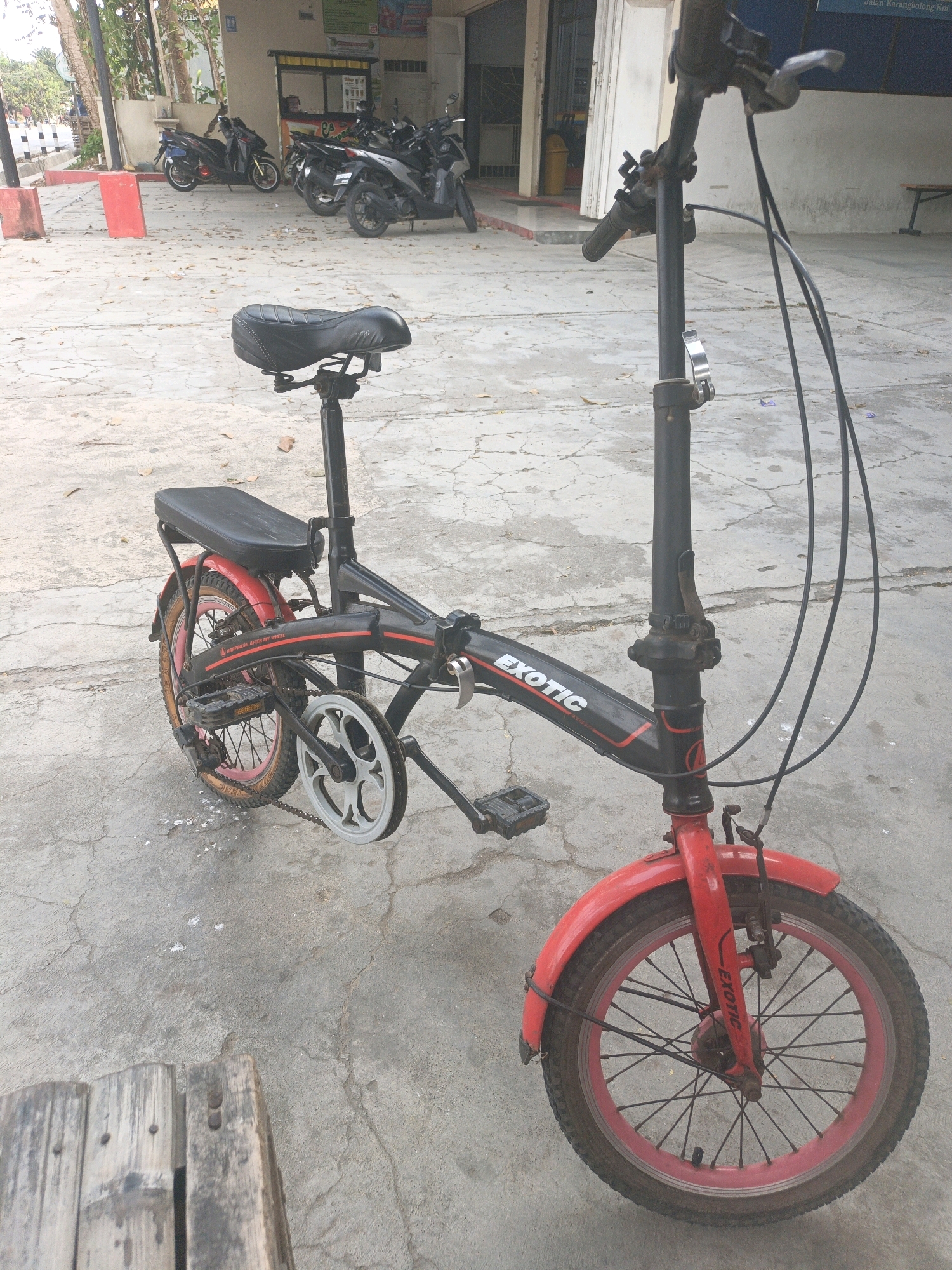 A red and black folding bicycle with the brand name "EXOTIC" visible on the frame is positioned in the foreground, angled towards the right. The bicycle has a black seat and black handlebars, with a silver quick-release lever on the steering column. The front wheel is red with black spokes, and the rear wheel is also red with black spokes. The bicycle is parked on a cracked concrete surface.
In the background, several motorcycles are parked in front of a building with a light-colored facade and an awning. The building appears to be a commercial establishment, possibly a shop or cafe. There are also trees and foliage visible, suggesting an outdoor setting. The scene is illuminated by natural daylight.
The location is specified as Buayan, Indonesia. The context suggests a public area, possibly a street or parking lot in a neighborhood. There are no people clearly visible interacting with the bicycle or the motorcycles. The overall impression is one of a quiet, ordinary day in an urban or semi-urban environment.
