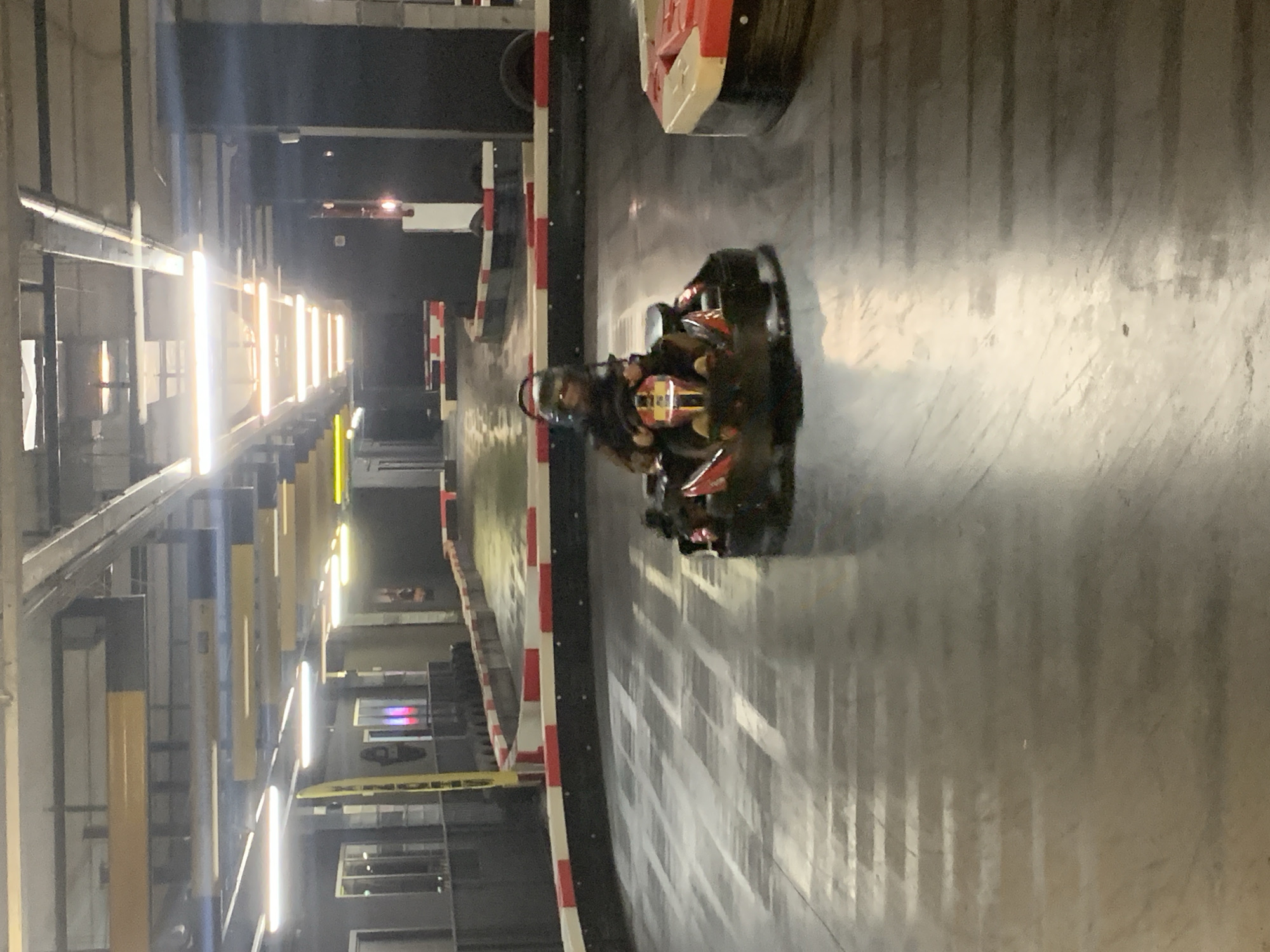 An indoor go-kart track at an unknown location features a person wearing a helmet driving a black and red go-kart. The track has a concrete surface with light and dark stripes, and is bordered by a barrier with red and white sections. Bright fluorescent lights illuminate the track from the ceiling.