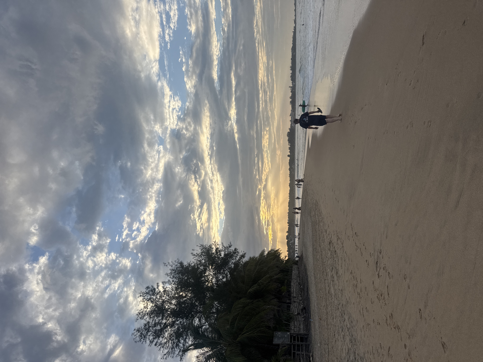 The image shows a beach scene at Arugam Bay, Sri Lanka. The sky is filled with dramatic clouds, with streaks of sunlight breaking through, suggesting either sunrise or sunset. The ocean is visible on the left, with gentle waves washing onto the shore. A sandy beach stretches out to the right, marked with footprints.
In the foreground, on the beach, a person is walking towards the water. They are wearing dark shorts and a dark t-shirt with white text, though it is difficult to read clearly. The person is holding a green object, possibly a surfboard or a paddleboard. Further down the beach, a line of people are visible walking along the shoreline, and some appear to be in the water. Lush green trees and palm fronds are seen on the left side of the image, adding to the tropical setting.
The overall mood is serene and peaceful, typical of a beach at this time of day. The lighting suggests a time of day with soft, warm light illuminating the clouds and the sand.