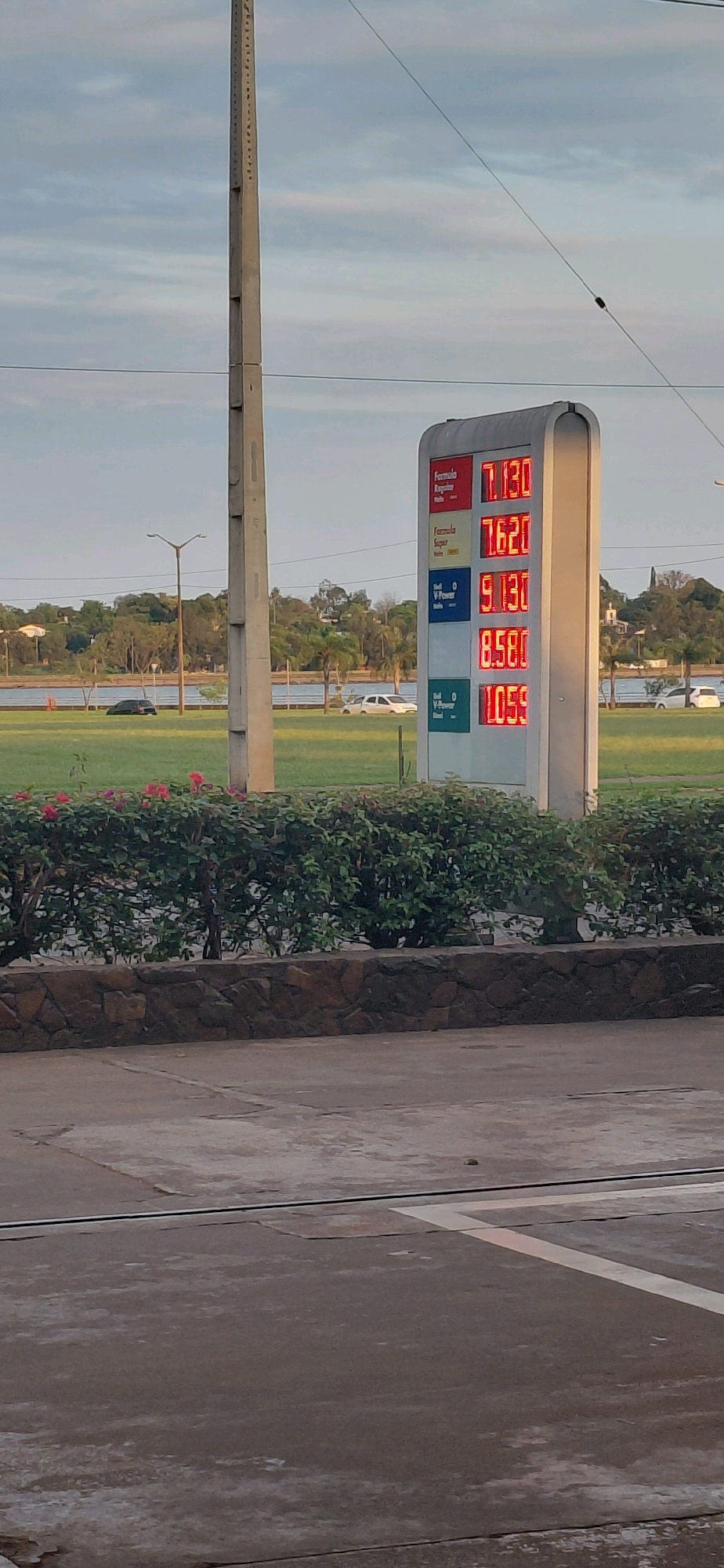 The image shows a gas station price board with red digital displays. The prices shown are 7.130, 7.620, 9.130, 8.580, and 1.059. Above the numbers, there are labels for different fuel types. The gas station appears to be located in Encarnacion, Paraguay, given the context of previous interactions. The background features a body of water, grassy areas, trees, and a cloudy sky. A concrete pole is visible to the left of the price board, and a row of green bushes with pink flowers is in the foreground. The ground is paved.