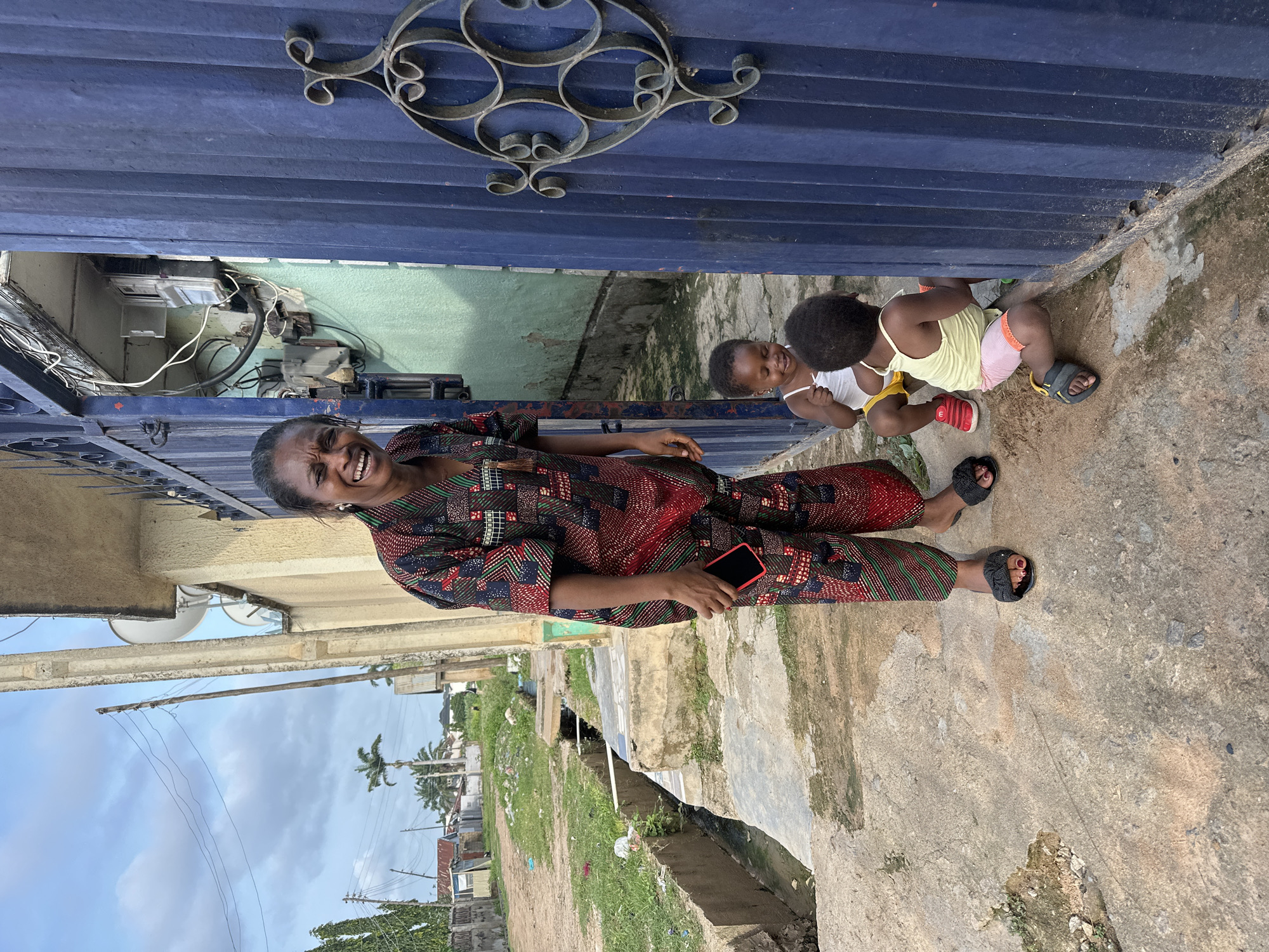 In Narayi, Nigeria, a woman in a vibrantly patterned, traditional outfit smiles broadly, holding a red smartphone. She stands on a concrete path next to a blue metal gate. Two young children are near the gate: one, a toddler, sits with a surprised expression, wearing a white tank top and yellow shorts, while another child, partially visible, crouches near the gate. The environment includes a drainage ditch to the left, a building with electrical meters and wiring visible behind the gate, and a street with houses and vegetation in the background under a cloudy sky.