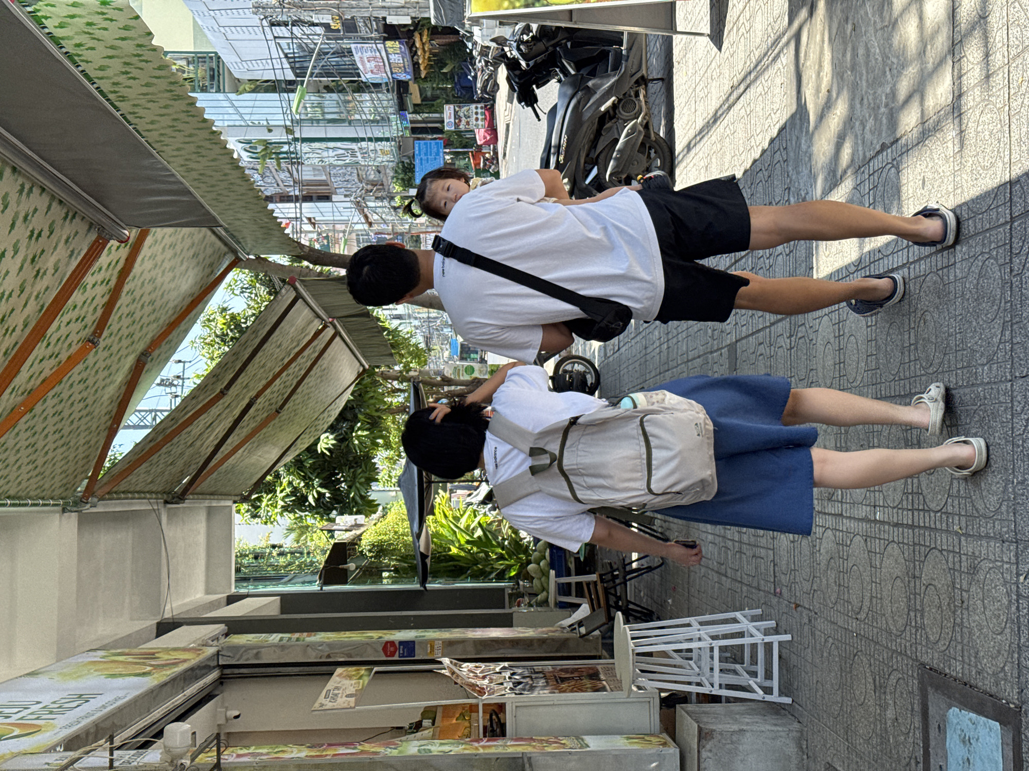 Two individuals and a child are walking on a tiled sidewalk in Nha Trang, Vietnam. The person in the foreground, viewed from behind, wears a white t-shirt, a light-colored backpack, and a blue skirt, and is walking on the right side of the sidewalk. To their right, a person viewed from behind wears a white t-shirt, black shorts, and is carrying a young child in their arms. The child is looking back at the camera. The sidewalk is lined with shops and awnings. To the left, there are tables and chairs outside a shop with signage featuring fruit. To the right, motorcycles are parked along the street. The environment suggests a commercial area with outdoor seating and pedestrian traffic.