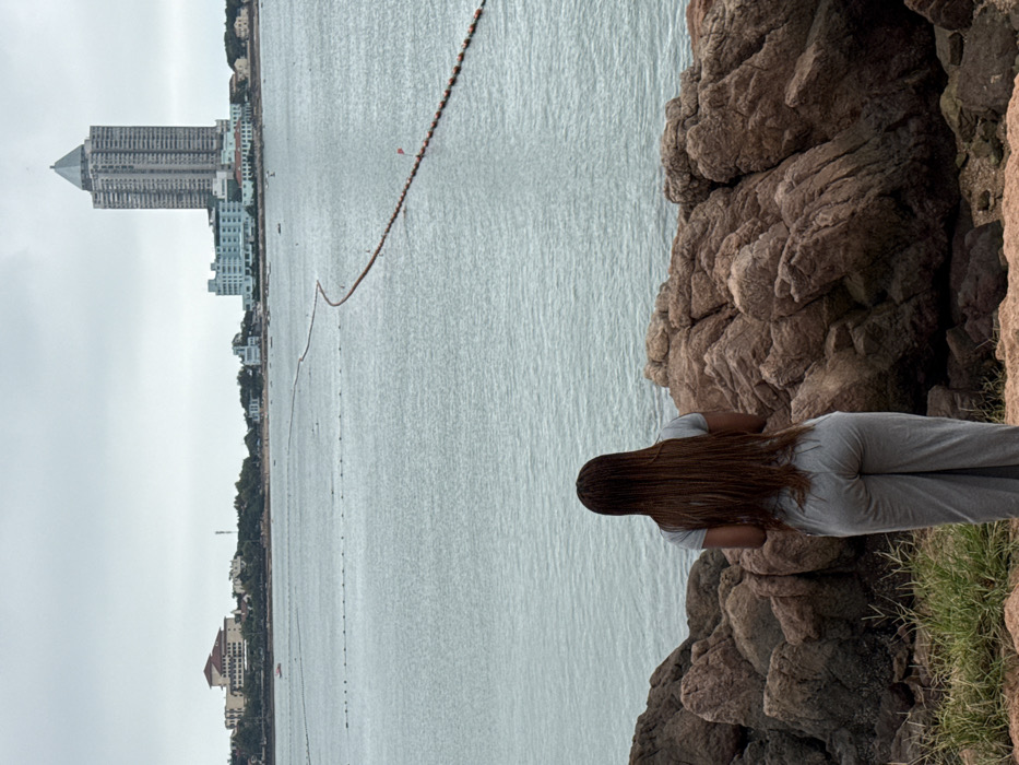 The image displays a coastal panorama within Shinan District, Qingdao, China, under an overcast, uniformly grey sky. In the immediate foreground, a person is seen from behind, standing on a rugged outcrop composed of reddish-brown rock formations and sparse green vegetation. The individual presents long, reddish-brown braided hair and wears a light grey short-sleeved t-shirt paired with grey loose-fitting pants. The person is oriented away from the camera, observing the scene across the water.
The midground features a calm, grey-blue bay. Two distinct lines of buoys are visible on the water; a closer, shorter line of dark buoys, and a longer, winding line of orange and white buoys marked with occasional small red flags extending towards the horizon. A small red paddle boat is discernible on the water near the distant shore.
The background reveals the urbanized coastline of Shinan District. Dominating the skyline is the tall, slender Qingdao International Financial Center, identifiable by its distinctive pointed, multi-faceted roof. Adjacent to it on the left stands a lower, light blue building complex. Further left, various other buildings, some featuring red-tiled roofs, are situated along the distant shore, with subtle indications of a beach area.