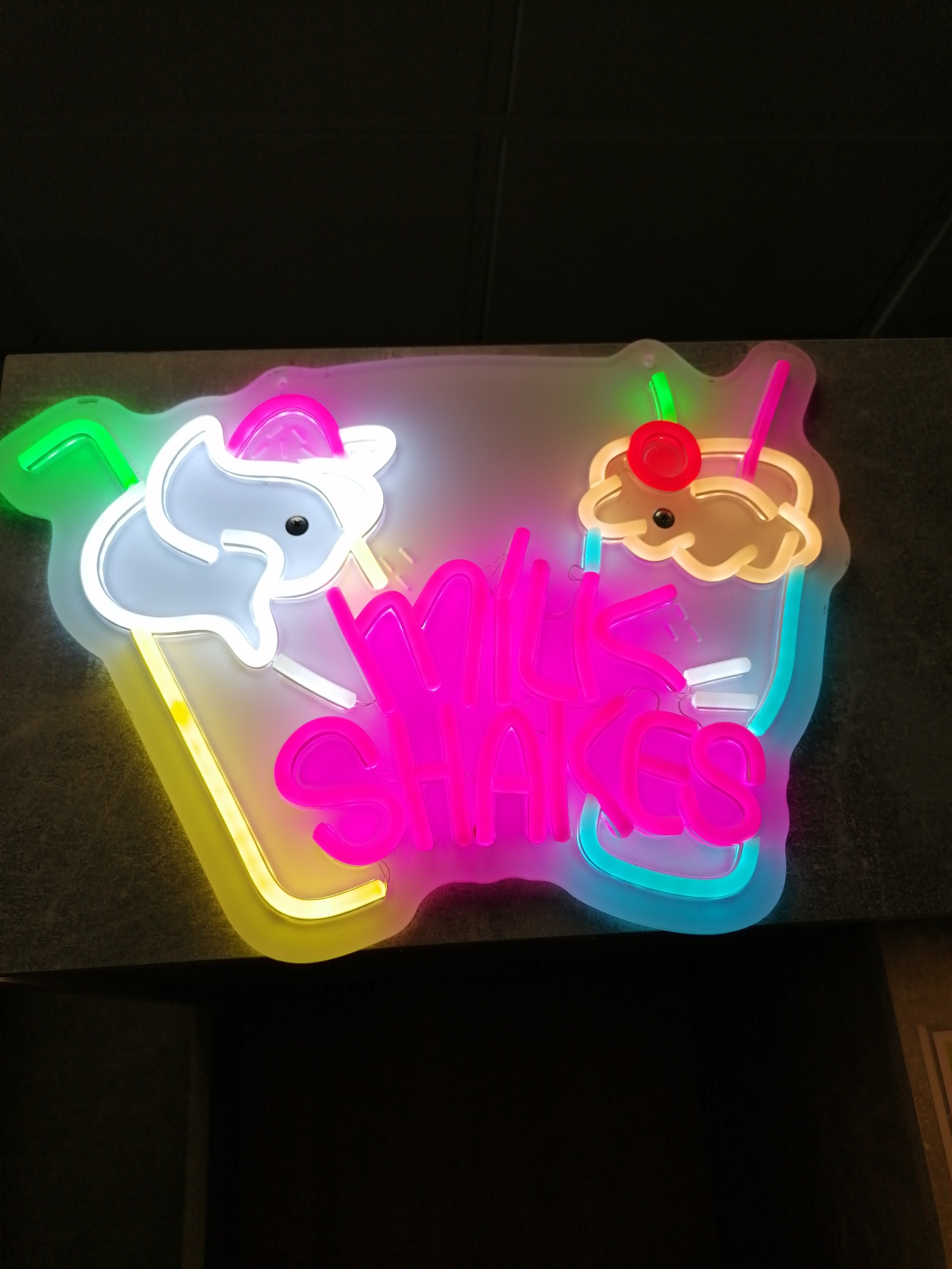 A neon sign, with two milkshake glasses and the words "MILK SHAKES" in pink neon, is mounted on a dark gray surface. The left milkshake is in a yellow glass with a white milkshake topped with a pink swirl, a green straw, and a small black circle. The right milkshake is in a blue glass and topped with a brown swirl and a red cherry. The setting appears to be indoors. The location is Unknown, Unknown.