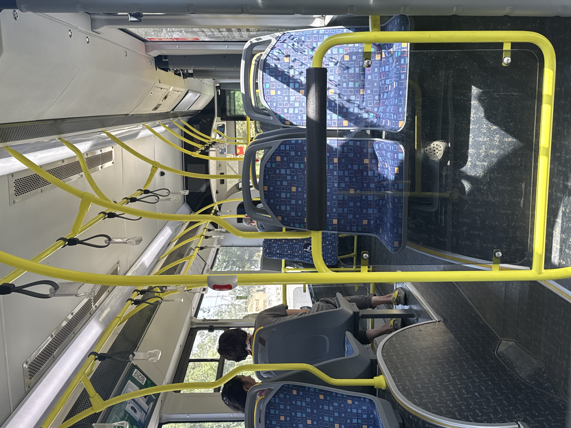 The image shows the interior of a bus, likely in Kashka Suu, Kyrgyzstan, with a focus on the seating area and yellow overhead handrails. Two passengers are visible in the foreground, seated and facing forward. One passenger, on the left, has dark hair and is wearing a pink shirt. The passenger to their right has short brown hair and is wearing a grey t-shirt and grey pants. Several rows of blue and patterned seats are visible throughout the bus. Yellow metal poles and handrails are prominent, providing support for standing passengers. The floor of the bus is dark and appears to be a textured material. Sunlight streams in through the windows, casting shadows on the seats and floor. The environment outside the windows suggests a bright, possibly warm day, with trees visible. There are also various interior bus fixtures, including vents and small red buttons, attached to the ceiling and side panels.