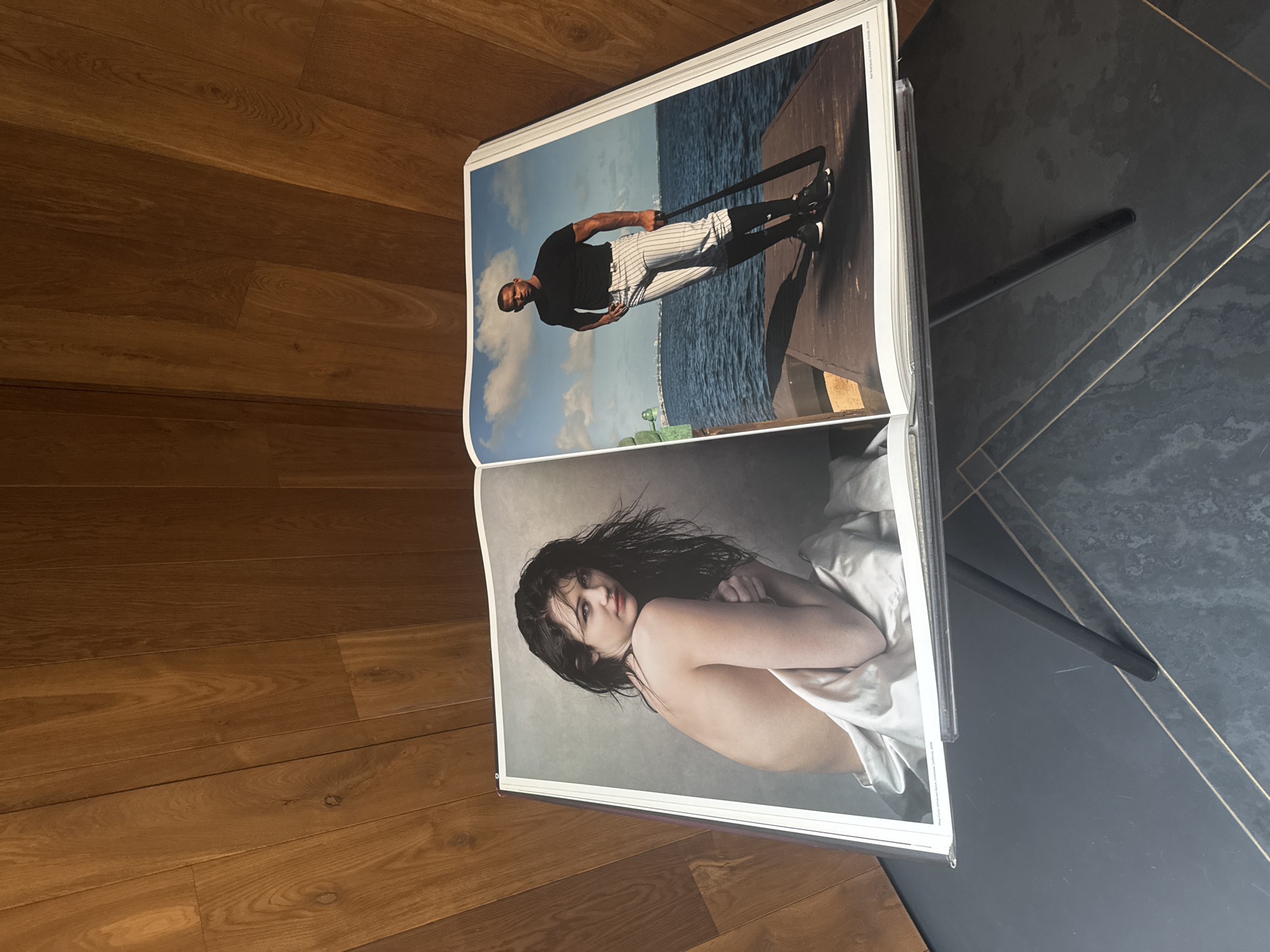 The image shows an open book or magazine propped up on a stand. The right page features a photograph of a man in athletic wear standing on a wooden pier or dock, with water and a city skyline in the background. The left page shows a photograph of a woman wrapped in a white sheet or blanket, exposing one shoulder. The stand is on a surface with a geometric pattern, possibly a tabletop or floor. To the left, there's a wooden floor with planks. The location in Mexico City, Mexico is not evident from the image.