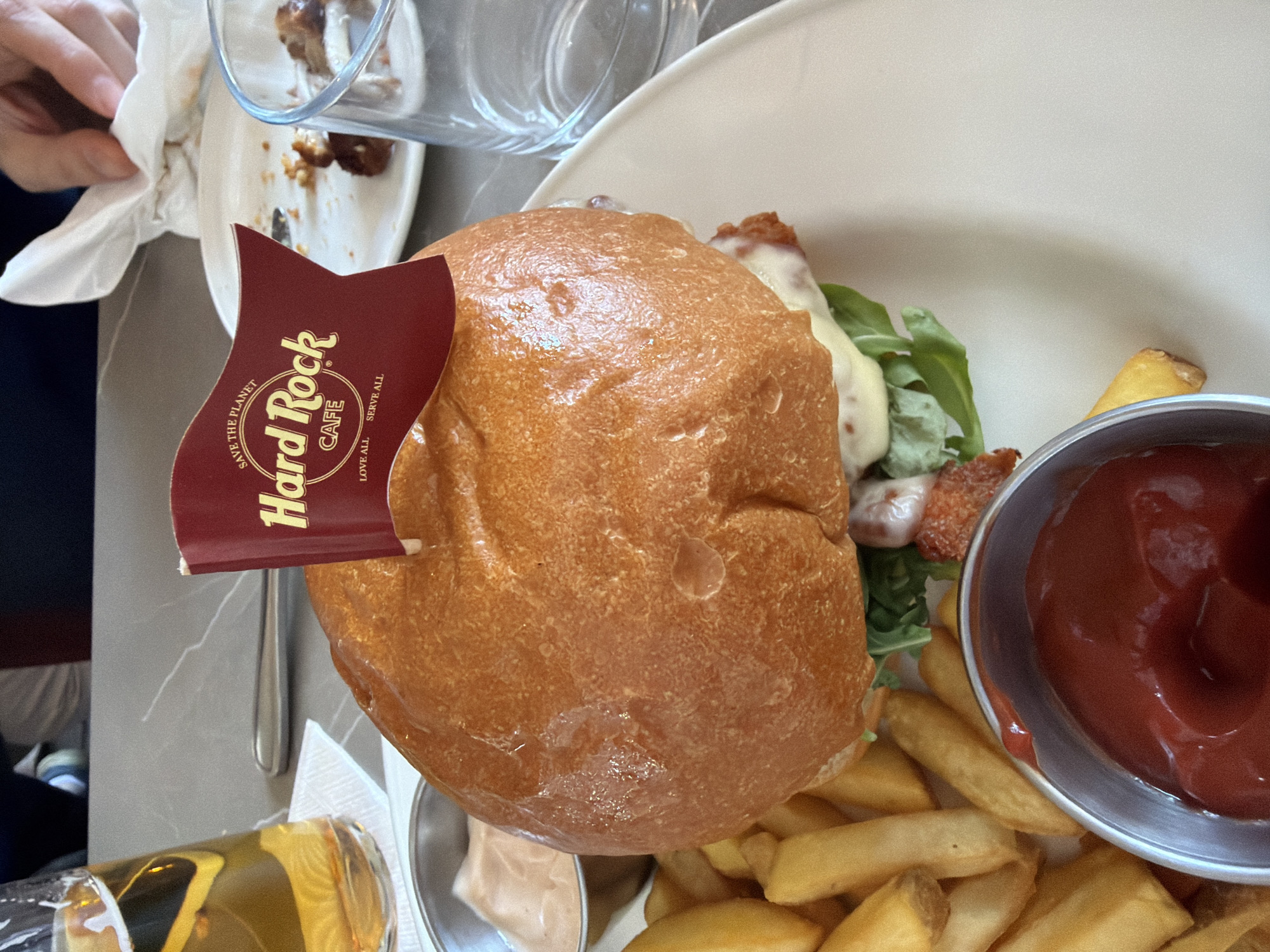 The image shows a partially eaten meal at the Hard Rock Cafe in Milan, Italy. A burger with a "Hard Rock Cafe" flag is the main focus, served on a white plate with fries and small metal bowls of ketchup and another sauce. A partially full beer glass is at the bottom left, while a used plate with leftover food scraps is at the top left, along with a person holding a napkin. The table is grey and textured, possibly marble or a similar material. A silver spoon is visible next to the burger.