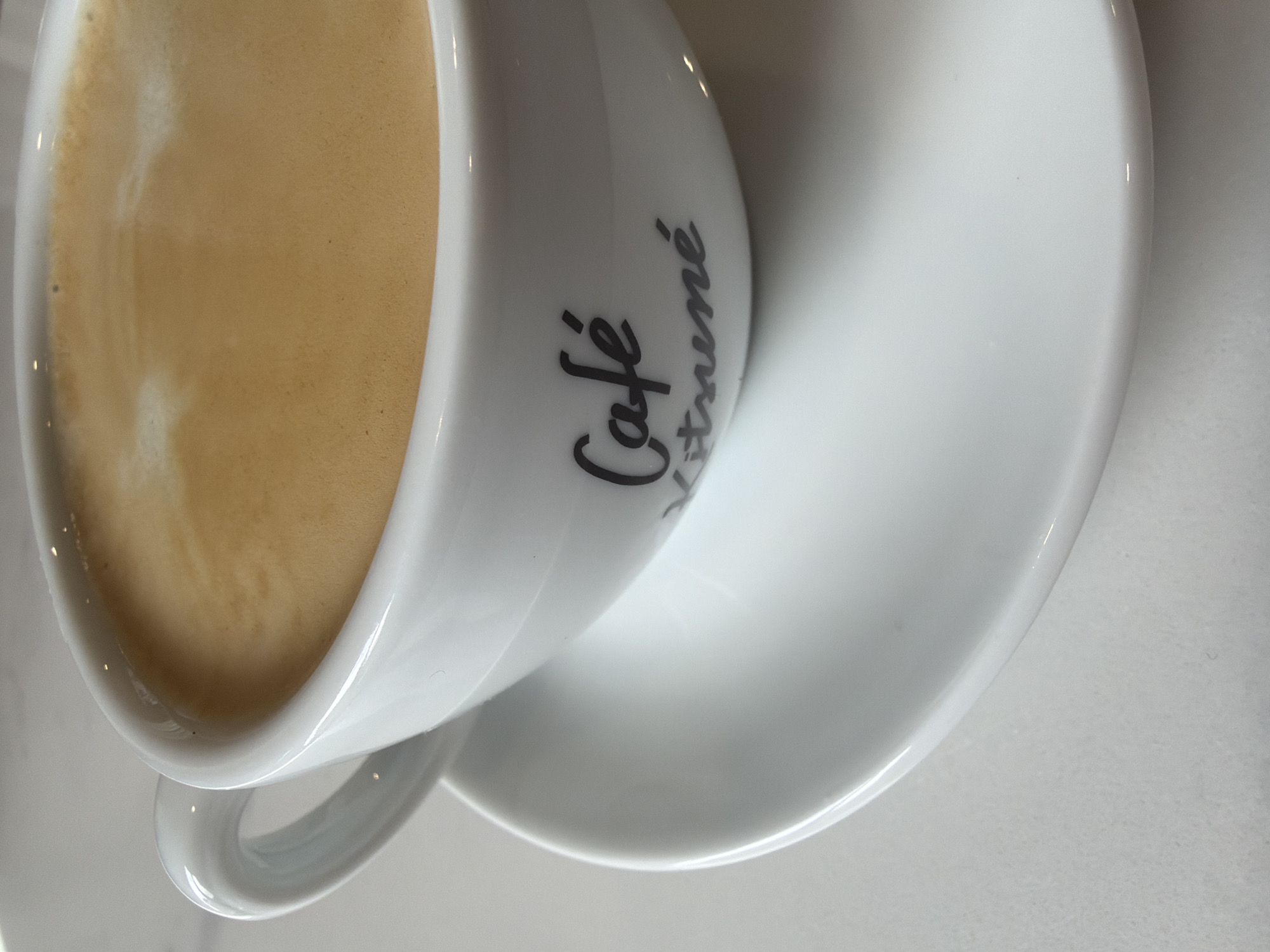 A close-up, high-angle shot depicts a white ceramic cup filled with a light brown, frothy beverage. The cup rests on a matching white saucer. The words "Café Kitsuné" are printed in black, stylized script on the side of the cup. The background is a light, neutral surface, possibly a table. The image contains no people or actions. The location in the city of Yerevan, Armenia cannot be determined based on visual information.
