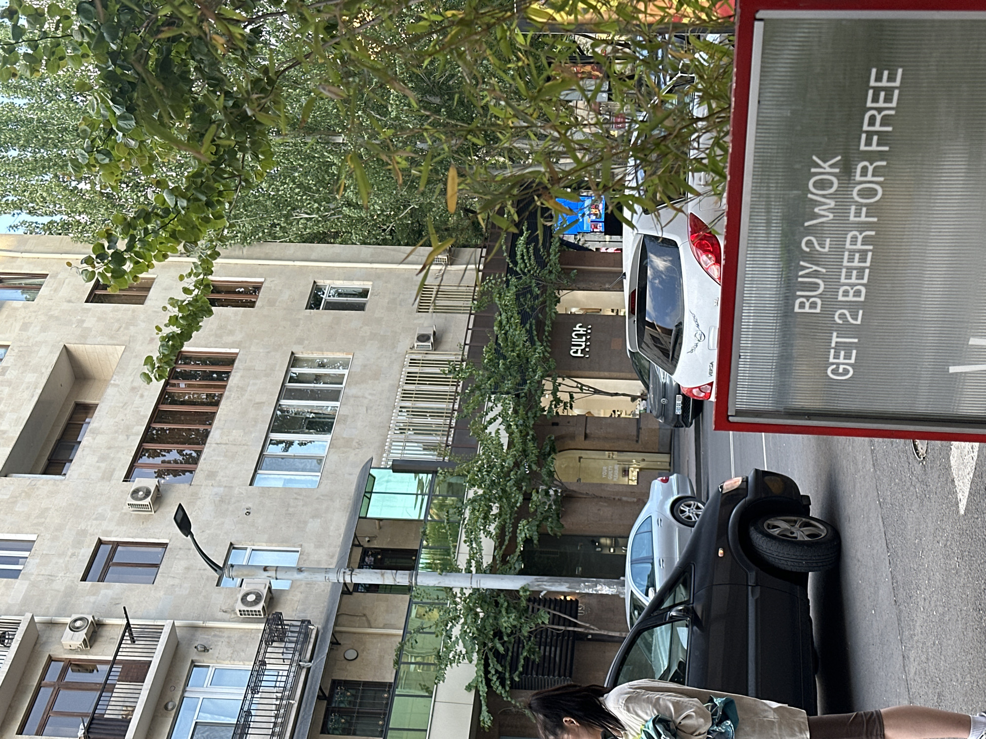 The scene is a street in Yerevan, Armenia, featuring an apartment building on the left. The building is light grey, with multiple windows, air conditioning units, and balconies. A black street light is visible in front of the building. On the right side of the image, there are two cars parked on the street, one black and one silver. A large sign with red trim advertises "BUY 2 WOK GET 2 BEER FOR FREE." In the foreground, a person wearing a beige shirt and brown shorts is partially visible. The environment also includes foliage from trees. There are also signs with writing in Armenian, including, "YOUR GUILTY PLEASURE" and "ԲԱԼԴԻ." The cars have license plates of "20101 (65)" and "SL VERSA".
