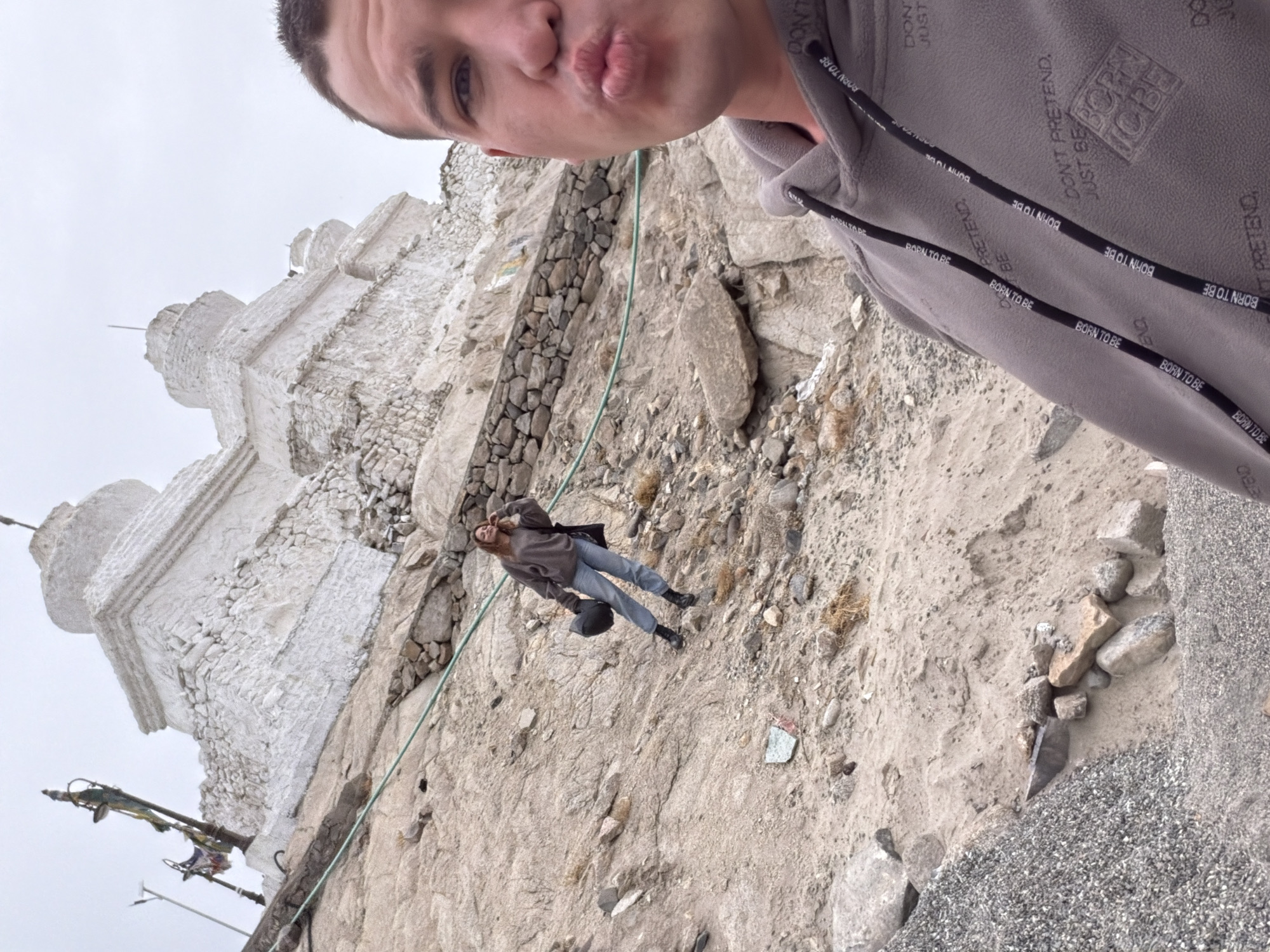 A man in the foreground is taking a selfie. He has short hair and is making a kissing face. He is wearing a gray hooded sweatshirt with "BORN TO BE" and "DON'T PRETEND" printed on it. Behind him, a woman is standing on a rocky hillside, holding a black helmet. She is wearing a gray sweatshirt and blue jeans. In the background, there is a large white stupa-like structure. The environment appears to be arid and mountainous, consistent with the location of Thiksey, India, where the Thiksey Monastery is located. The stupa is a prominent feature of the monastery. The ground is covered in sand, gravel, and rocks.