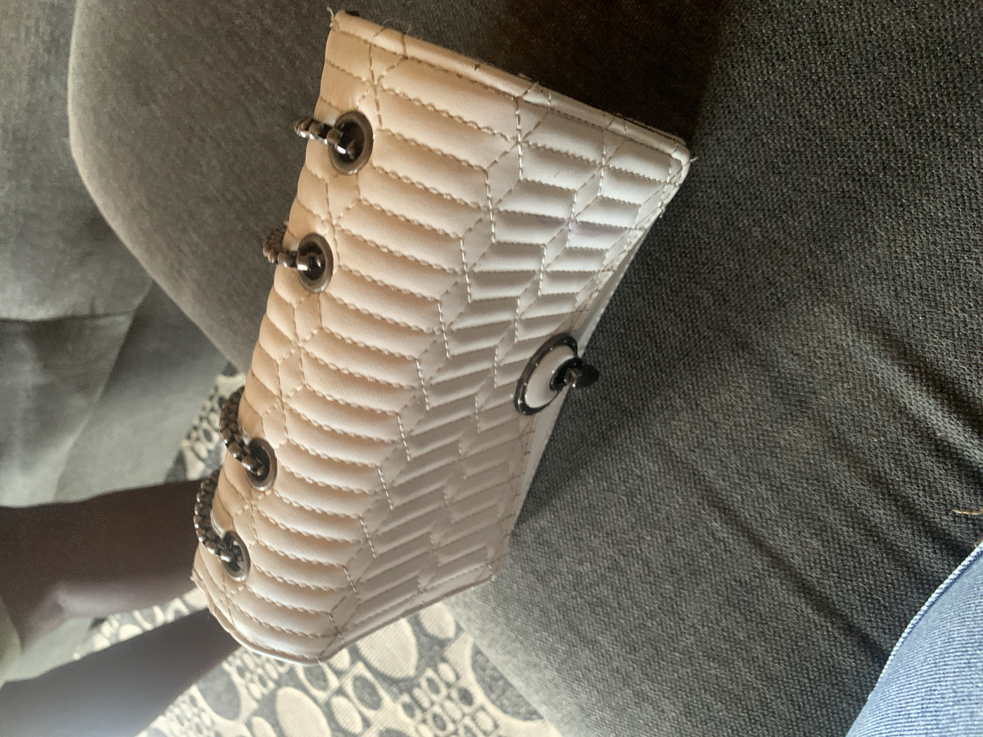 A close-up, overhead shot shows a white, quilted, rectangular handbag with silver hardware. The handbag is resting on a dark gray, textured surface, likely clothing or upholstery. The background is blurred but appears to be a patterned fabric and a person's legs. A faint purple mark is visible on the front of the handbag. The image does not contain any information that would allow for its location in Sabon Tasha, Nigeria.