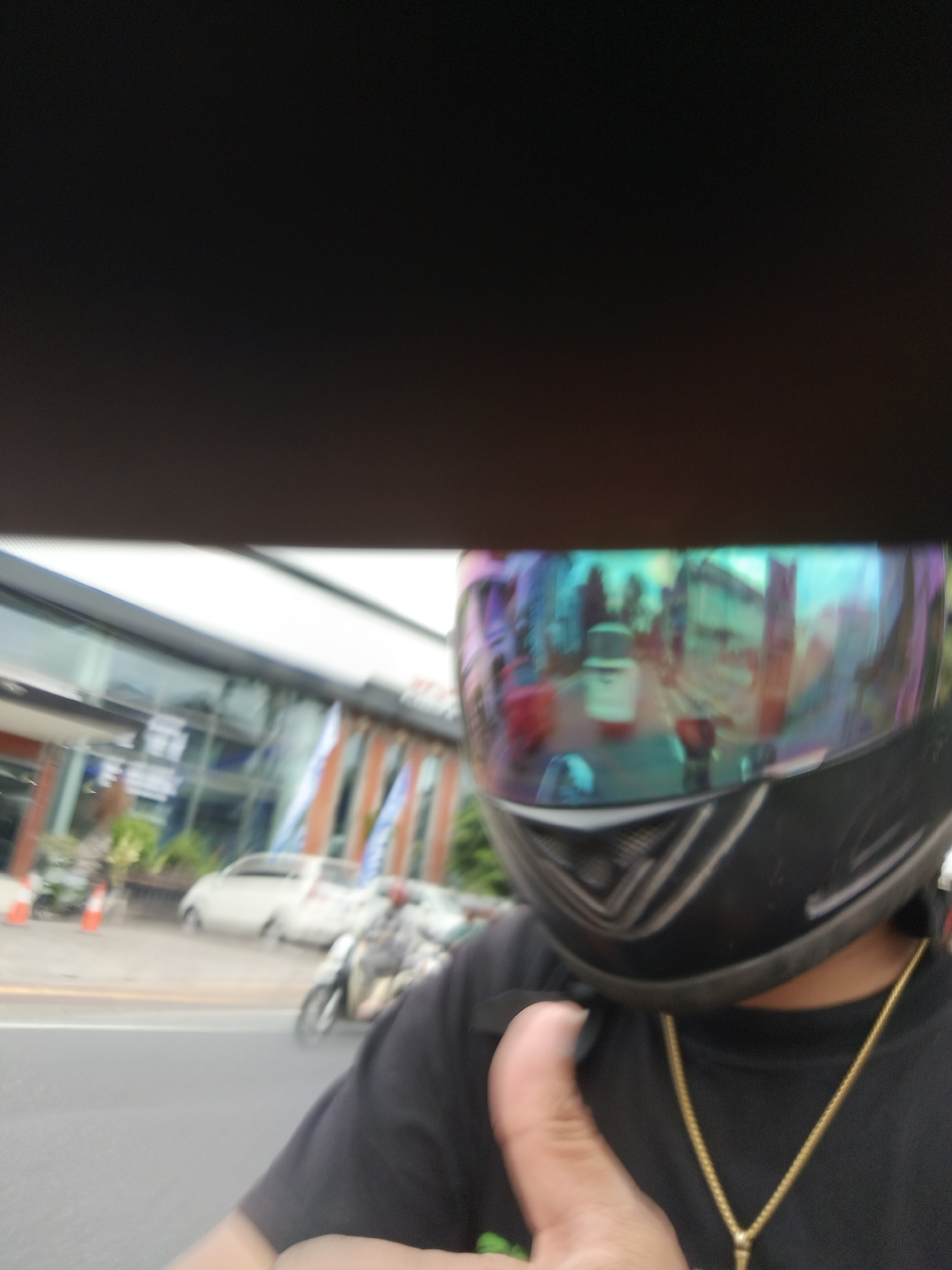 The image shows a first-person perspective of someone wearing a black motorcycle helmet with a reflective visor. The visor reflects a street scene with buildings, other vehicles, and possibly people, all rendered in a distorted and colorful manner. In the foreground, the person's thumb is visible, giving a thumbs-up gesture. The person is wearing a black t-shirt and a gold chain. Behind the person, a building with large glass windows and some orange striped flags can be seen. Traffic cones and parked cars are also visible on the side of the road. The overall impression is of a person on a motorcycle, possibly at a traffic light or stopped briefly. The environment suggests an urban setting, likely in Denpasar, Indonesia, given the context provided.