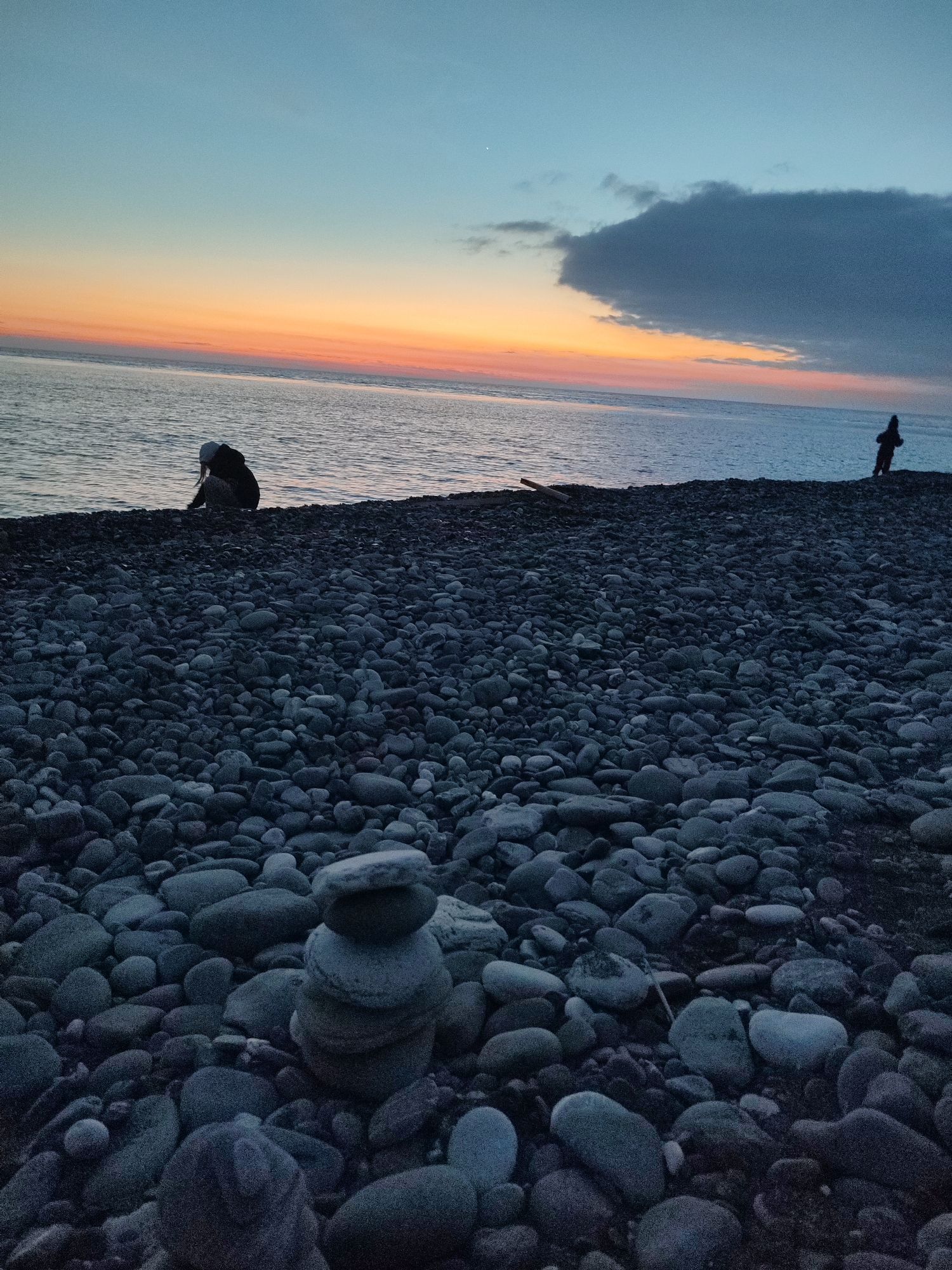 The image depicts a pebble beach in Sochi, Russia, at twilight. The sky transitions from light blue to orange near the horizon. A dark cloud rests near the horizon on the right side of the image. The sea is calm. A stack of flat, grey pebbles stands in the foreground. To the left, a person kneels near the water's edge. To the right, a second individual stands further down the beach. A piece of driftwood lies on the beach between them. The scene is composed mainly of the pebble beach, sea, and sky.