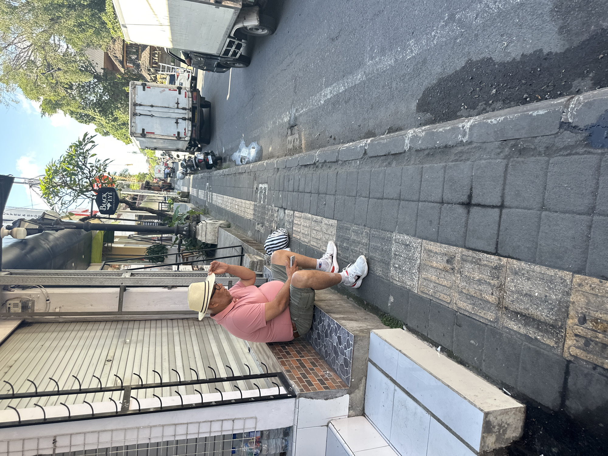 A man wearing a pink polo shirt, green shorts, white socks, and white sneakers sits on a tiled bench in front of a closed storefront on a sidewalk in Ubud District, Indonesia. He has a straw hat on his head and is looking down at his phone, which he holds in his right hand. His left hand is raised to his hat. A black and white striped bag rests on the ground next to him. The sidewalk is paved with gray cobblestones and textured tiles for the visually impaired. Across the street, vehicles, including a white truck and motorcycles, are on the road. The environment appears to be a commercial street with trees and buildings lining the road. The sky is blue with scattered clouds.