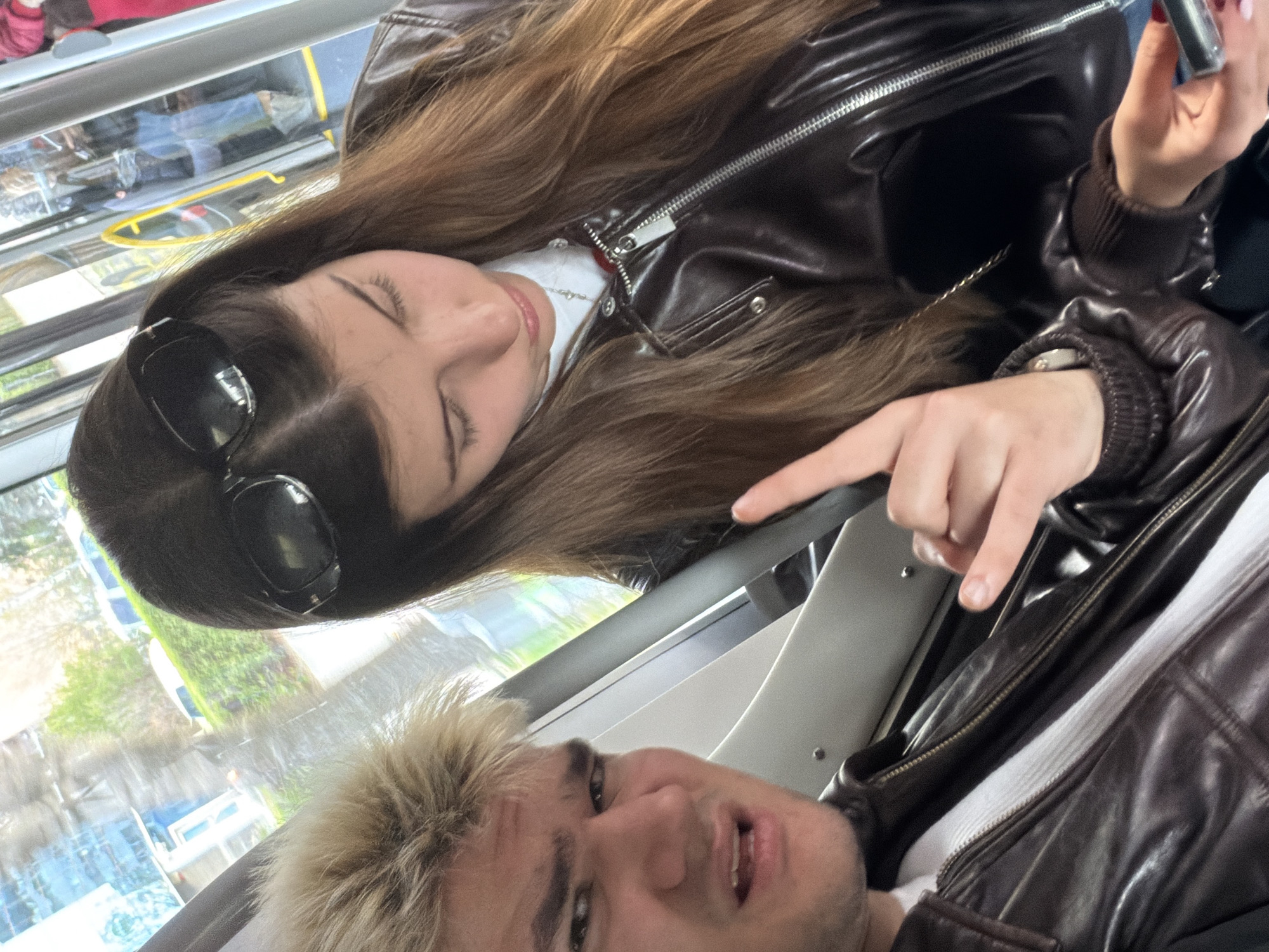 A young woman with long brown hair, wearing sunglasses and a brown leather jacket, is seated next to a young man with light blonde hair, also wearing a brown leather jacket. The woman's eyes are closed and she is holding a phone. The man is looking directly at the camera. The scene appears to be inside a public transportation vehicle, likely a tram or bus, in Sofia, Bulgaria, given the view from the window showing urban elements.