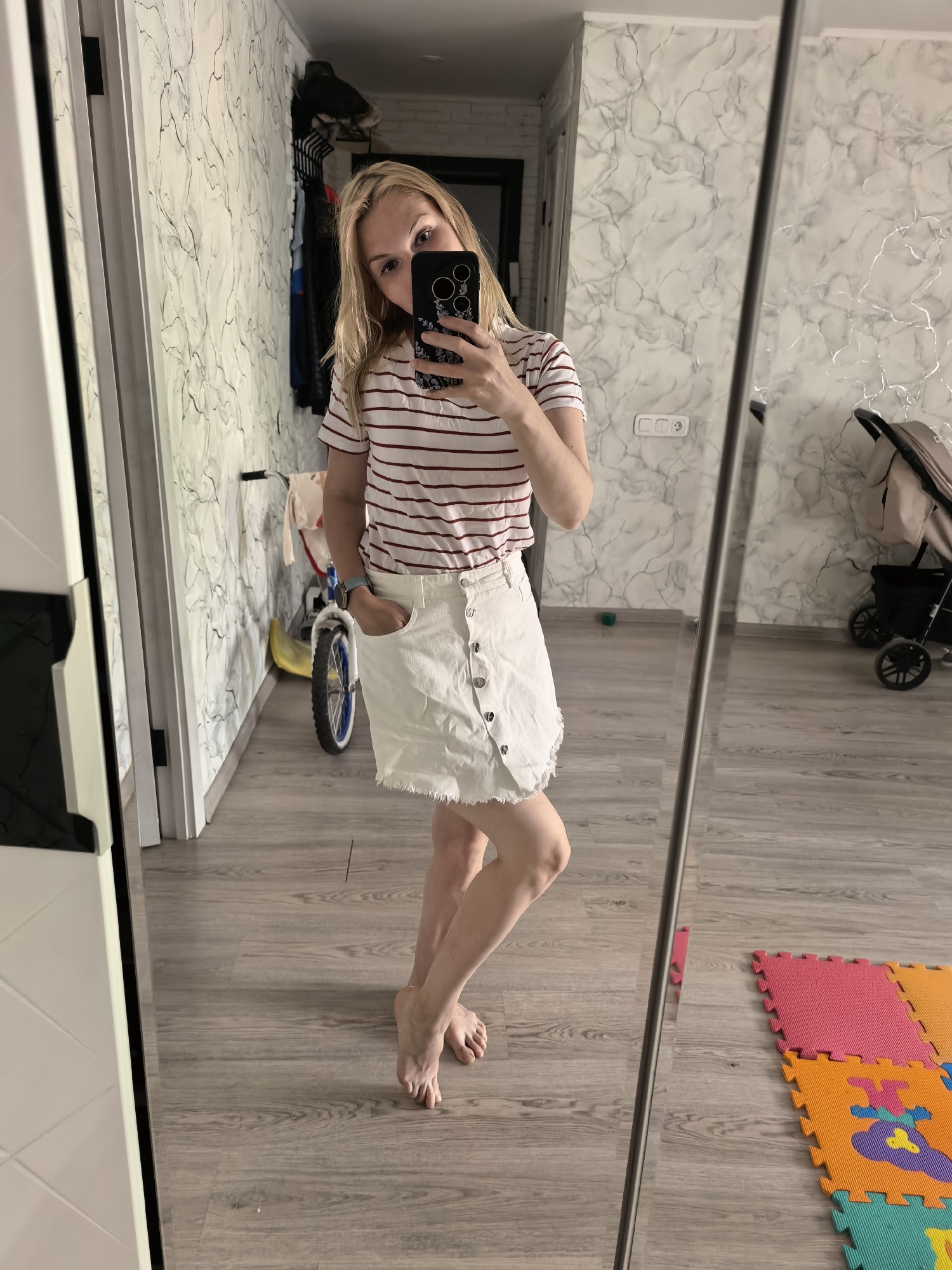 A woman with blonde hair is taking a mirror selfie indoors. She is wearing a red and white striped t-shirt and a white denim skirt with buttons. She is holding a black smartphone up to her face, partially obscuring it. The background includes a hallway with a marble-patterned wall, a child's bicycle, a baby stroller, and foam play mats on the floor. The flooring appears to be laminate or wood. There is also a door visible in the background, and a cabinet to the left of the mirror.