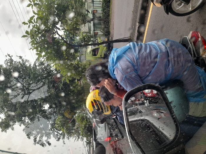 The scene depicts a street in Calukan RT 1 RW 5 Keboansikep, Indonesia, during rainfall, evidenced by numerous raindrops on the viewing surface and a passenger's attire. A motorcycle occupies the central foreground, carrying two individuals. The driver wears a yellow helmet with black stripes and partial text "max". The passenger, seated behind the driver, is clad in a translucent light blue poncho-style raincoat, with dark, wet hair; her left hand is raised near her ear.
In the lower-left, a rearview mirror reflects a sequence of vehicles, including a red scooter and a white car, indicating traffic behind the vantage point. Behind the central motorcycle, a dark car with a partially visible license plate is present. The far-right edge of the image shows the front wheel and engine component of a black motorcycle.
The background is characterized by an abundance of dense green trees under an overcast sky. A light green utility vehicle is discernible in the middle ground, partially obscured by foliage and a white fence structure. The road surface is asphalt, delineated by a yellow line and a raised curb. The overall environment suggests an urban or semi-urban setting experiencing active precipitation and vehicular movement.