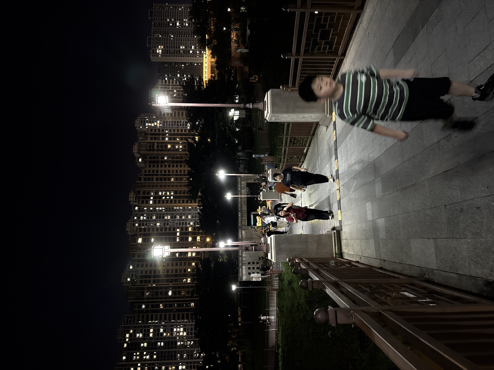 An outdoor night scene in Huizhou, China, shows a paved walkway with people walking. In the foreground, a young boy wearing a striped t-shirt and black shorts runs towards the camera. Behind him, several adults are walking away from the camera. A decorative metal railing runs along the side of the walkway. In the background, tall apartment buildings with illuminated windows are visible, suggesting a residential area within the city. Streetlights illuminate the walkway, casting shadows and creating a moderately lit environment. Trees line the area between the walkway and the apartment buildings. The overall atmosphere suggests a public park or communal space in a densely populated urban area at night.
