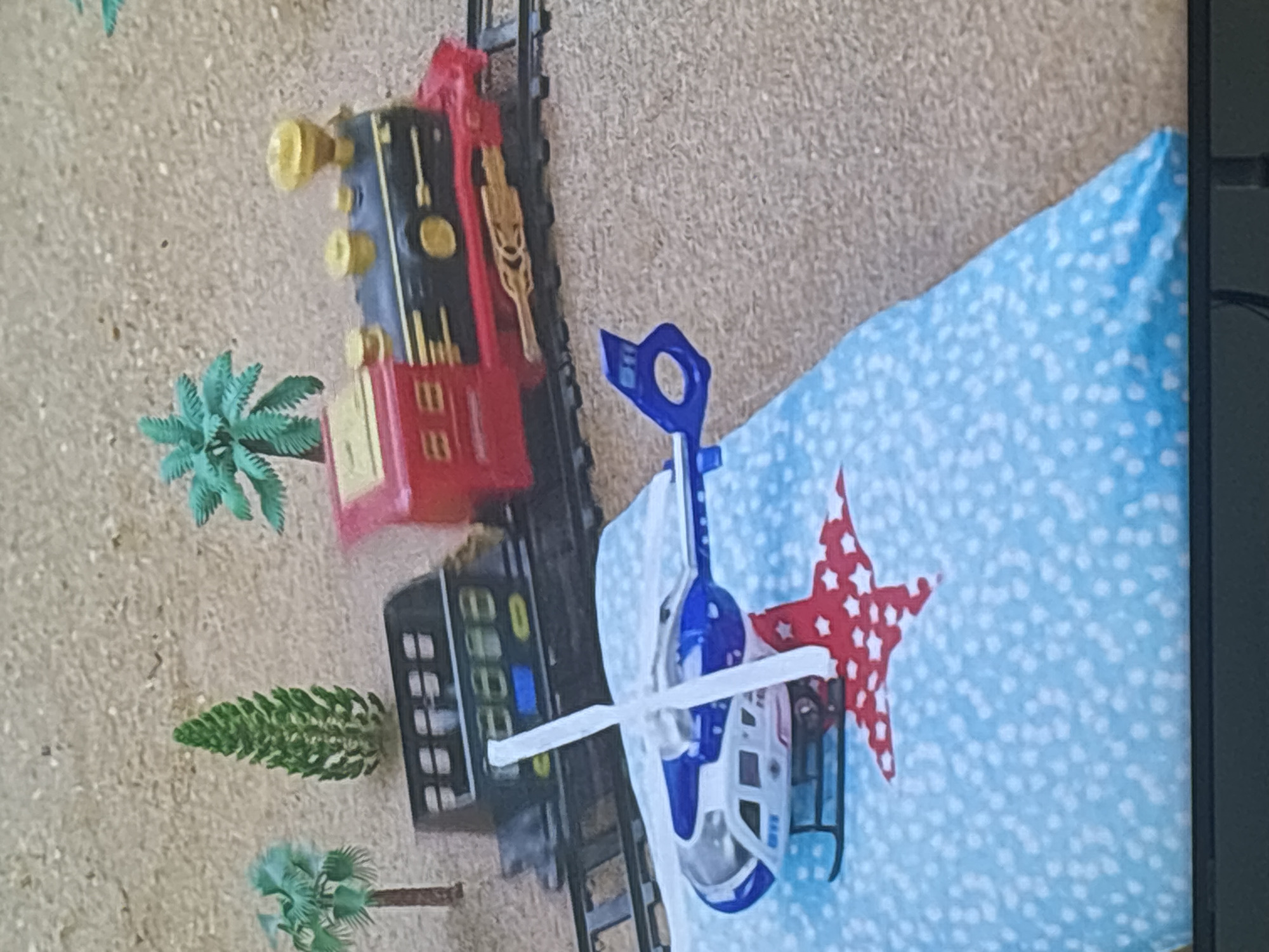 The image shows a toy helicopter and a toy train on a textured surface resembling sand. The helicopter is blue and white with a red star design beneath it. The train is red and black with gold accents and is on a track. There are miniature trees in the background. The scene appears to be indoors, possibly a child's play area, and there is no indication of the city of Dubai or the United Arab Emirates.