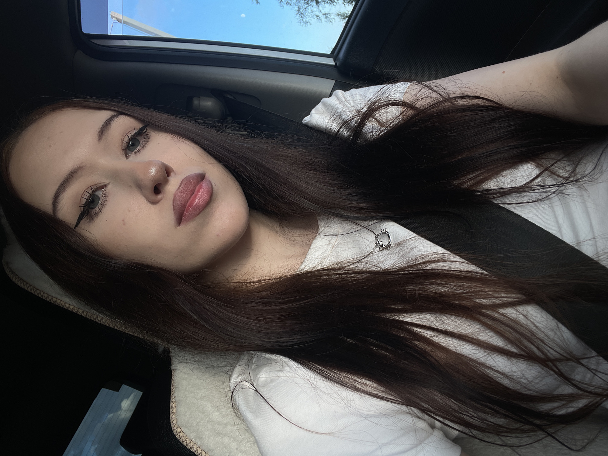 A young woman is captured in a close-up, selfie-style shot, taken from a slightly elevated angle. She has long, dark brown hair that cascades over her shoulders and chest. Her makeup is noticeable, with prominent eyeliner and full lips. She is wearing a white t-shirt, and a silver necklace with a pendant is visible. A black seatbelt is fastened across her body, suggesting she is in a vehicle.
The setting appears to be the interior of a car, with a glimpse of the sky and trees through an open sunroof. The light suggests it is daytime, possibly late afternoon, given the angle of the sun. The overall mood conveyed is relaxed and casual. No text is visible in the image. The location context provided is Gavrilovka, Russia.