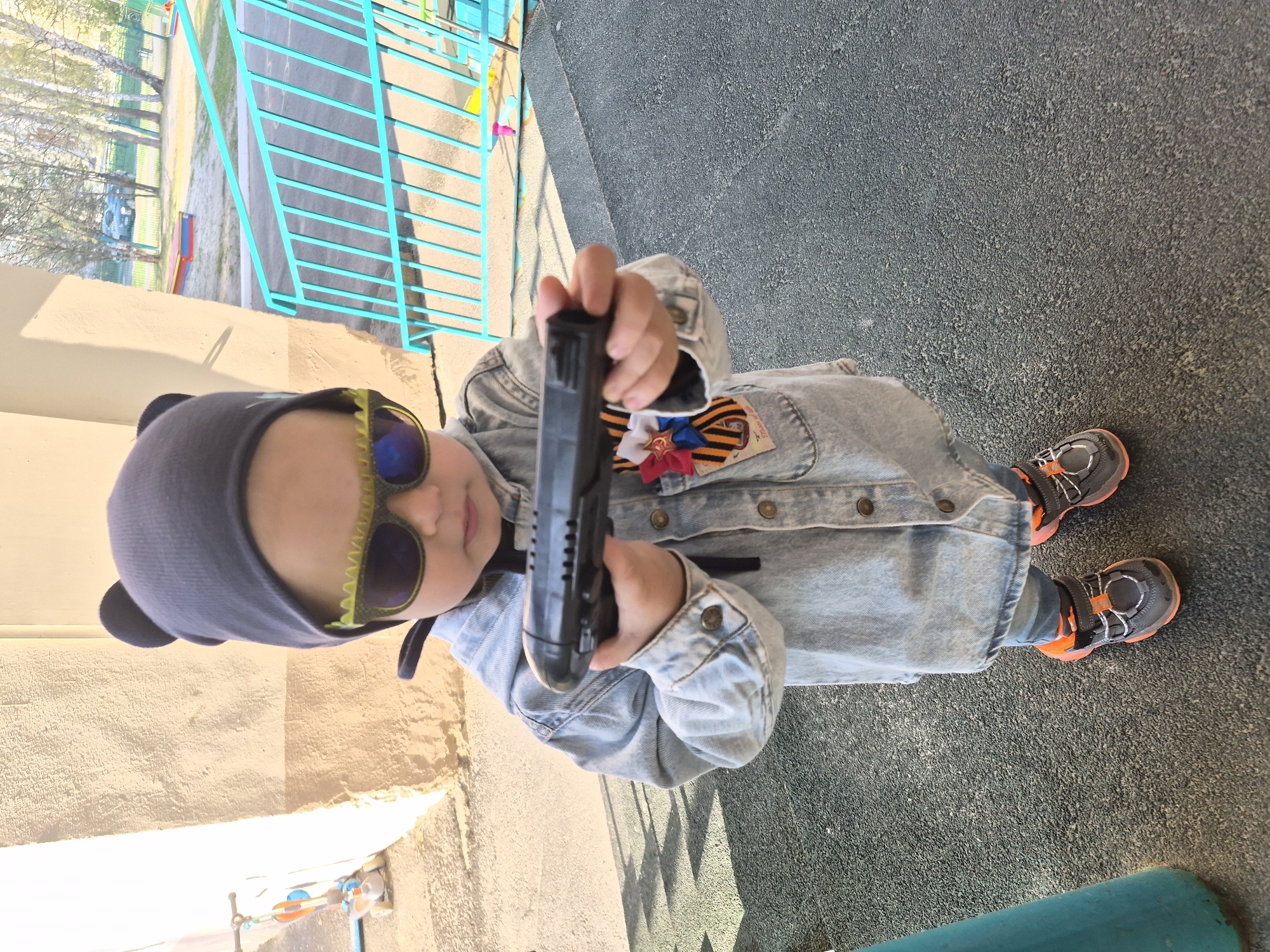 A child, wearing a blue cap with bear ears, green sunglasses, and a denim jacket with a St. George ribbon and a rosette of the Russian flag pinned to it, stands on a textured gray surface and holds a black object resembling a toy pistol in front of them. The child wears gray and orange sneakers. In the background, there is a low, light-colored building adjacent to a blue metal fence. Beyond the fence is a view of trees and playground equipment. This location is likely in Tobolsk, Russia, given the St. George ribbon and Russian flag rosette.