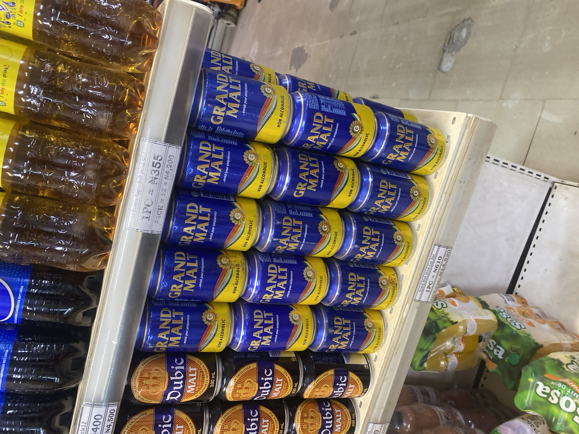The image shows a shelf in a Nigerian supermarket, likely located in Ikorodu, Nigeria, displaying various beverages. The foreground features stacks of canned drinks, primarily "Grand Malt" and "Dubic Malt," identifiable by their blue and yellow packaging. Some bottles of what appear to be soft drinks are also visible on the shelf above, along with other packaged beverages like "Sosa" juice drinks on a lower shelf. Price tags are visible on the shelves, indicating prices in Nigerian Naira (₦). The environment appears to be a retail setting with fluorescent lighting and tiled flooring. There are no people clearly visible in the image.