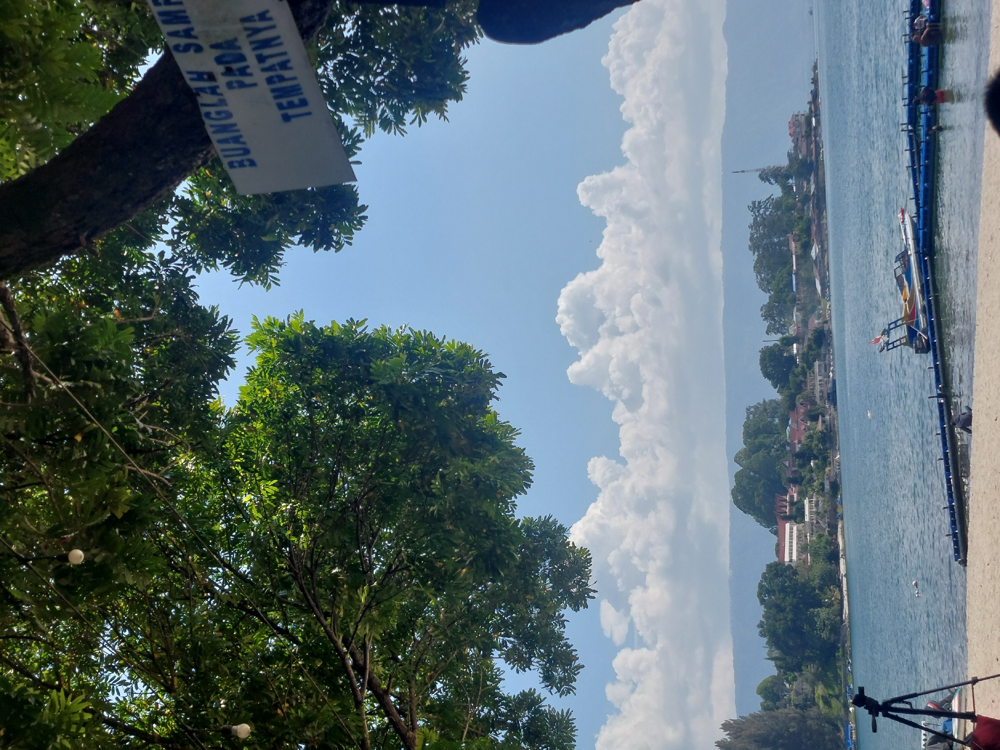 The image depicts a scenic view of a lakefront, likely in Parapat, Indonesia, on a clear day. In the foreground, a sandy beach is visible with a black tripod and a red bag. A blue and white floating dock extends into the water, where several people are seen swimming and interacting. A small boat is docked at the floating structure. The lake itself is calm and blue, with a distant shoreline featuring lush green trees and buildings, including what appears to be a resort or hotel. The sky is a bright blue, adorned with fluffy white clouds. Overhead, branches of a large tree frame the upper portion of the image, with two hanging light bulbs visible. A sign attached to the tree trunk reads "BUANGLAH SAMPA" which translates to "Dispose of trash". The overall atmosphere is tranquil and evokes a sense of leisure and natural beauty.