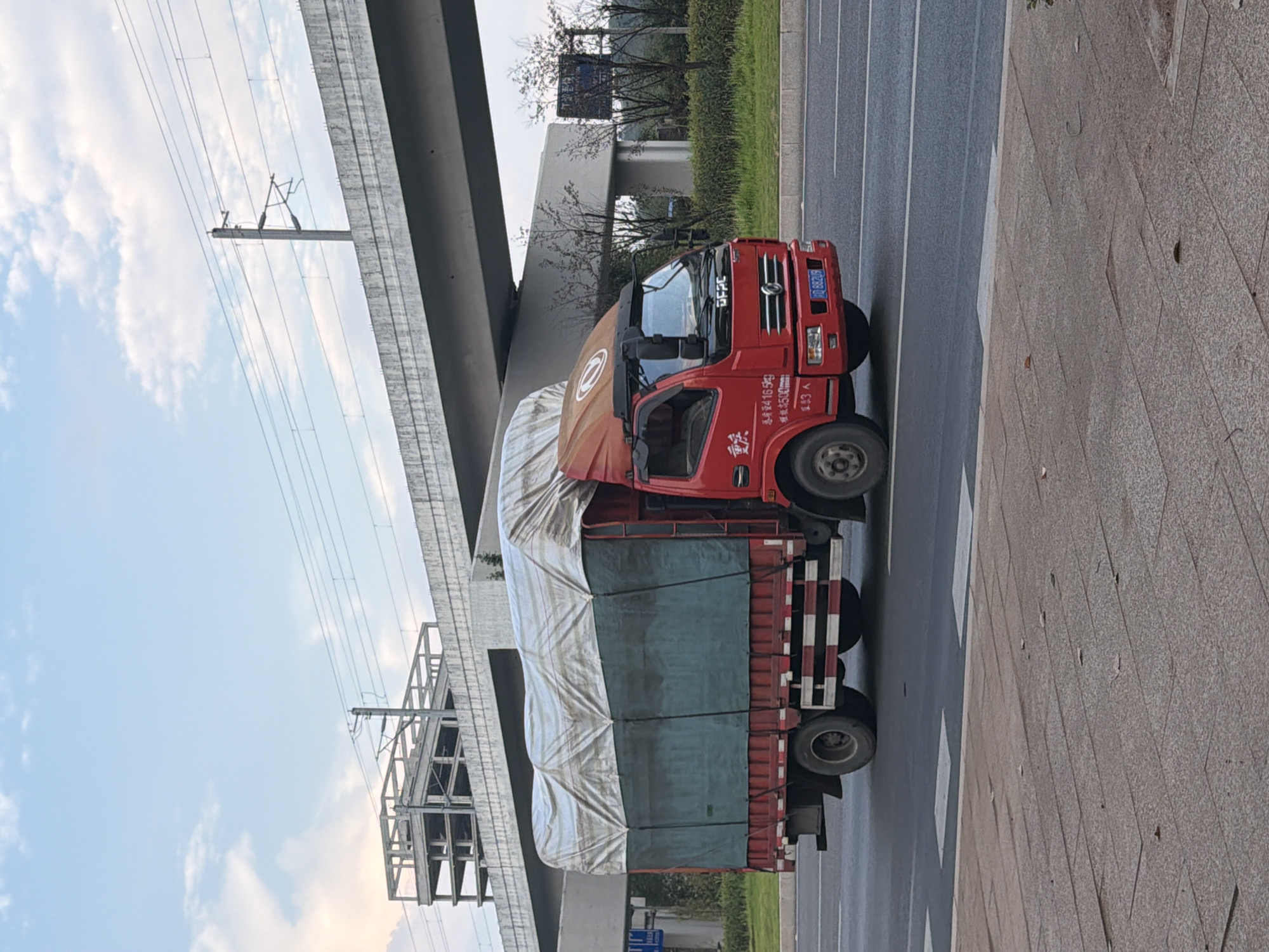 An outdoor, eye-level, medium shot shows a red, covered truck traveling on a road, with a concrete overpass and an unfinished building in the background. The truck is traveling from left to right, and its cargo is covered with a tarp. The truck's license plate reads "BD 88208". The sky is blue with some clouds. The image appears to be taken in Jiangjin, China, as suggested by the Chinese characters on the truck and a sign in the background, though the specific location within Jiangjin is not discernable.