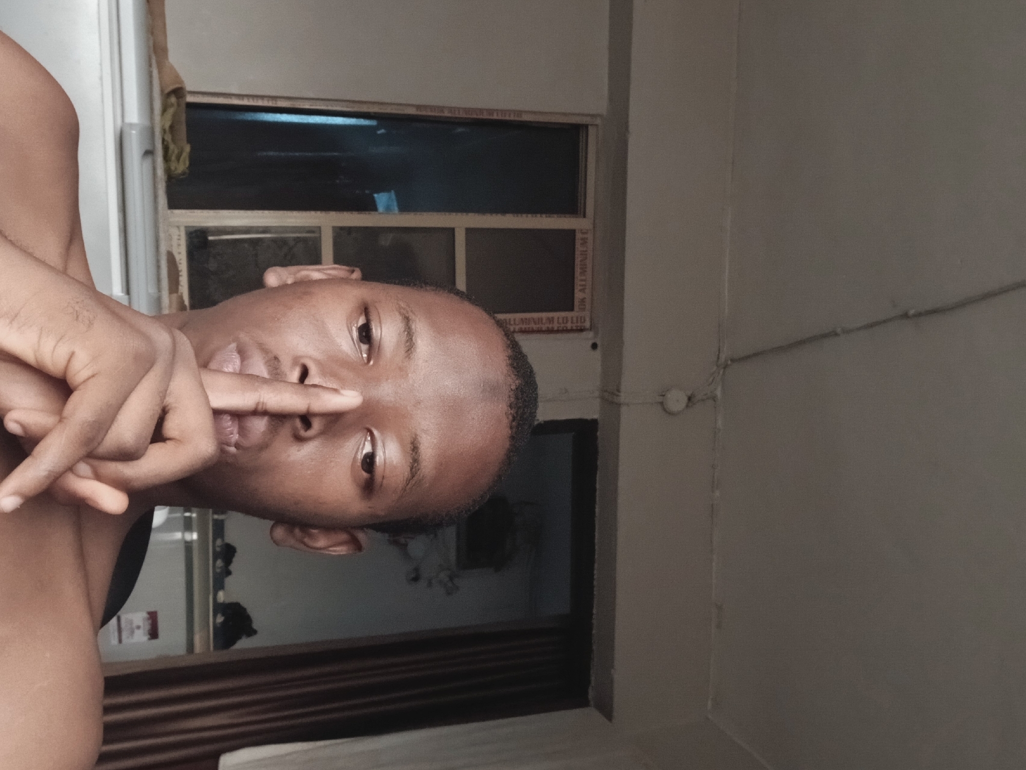 A man with dark skin and short hair is posing for a photo with his finger on his nose. He is indoors, possibly in Lagos, Nigeria, based on the aluminum window frame visible in the background. The lighting is dim. The background includes a window with a grid pattern and a beige-colored wall with electrical wiring.